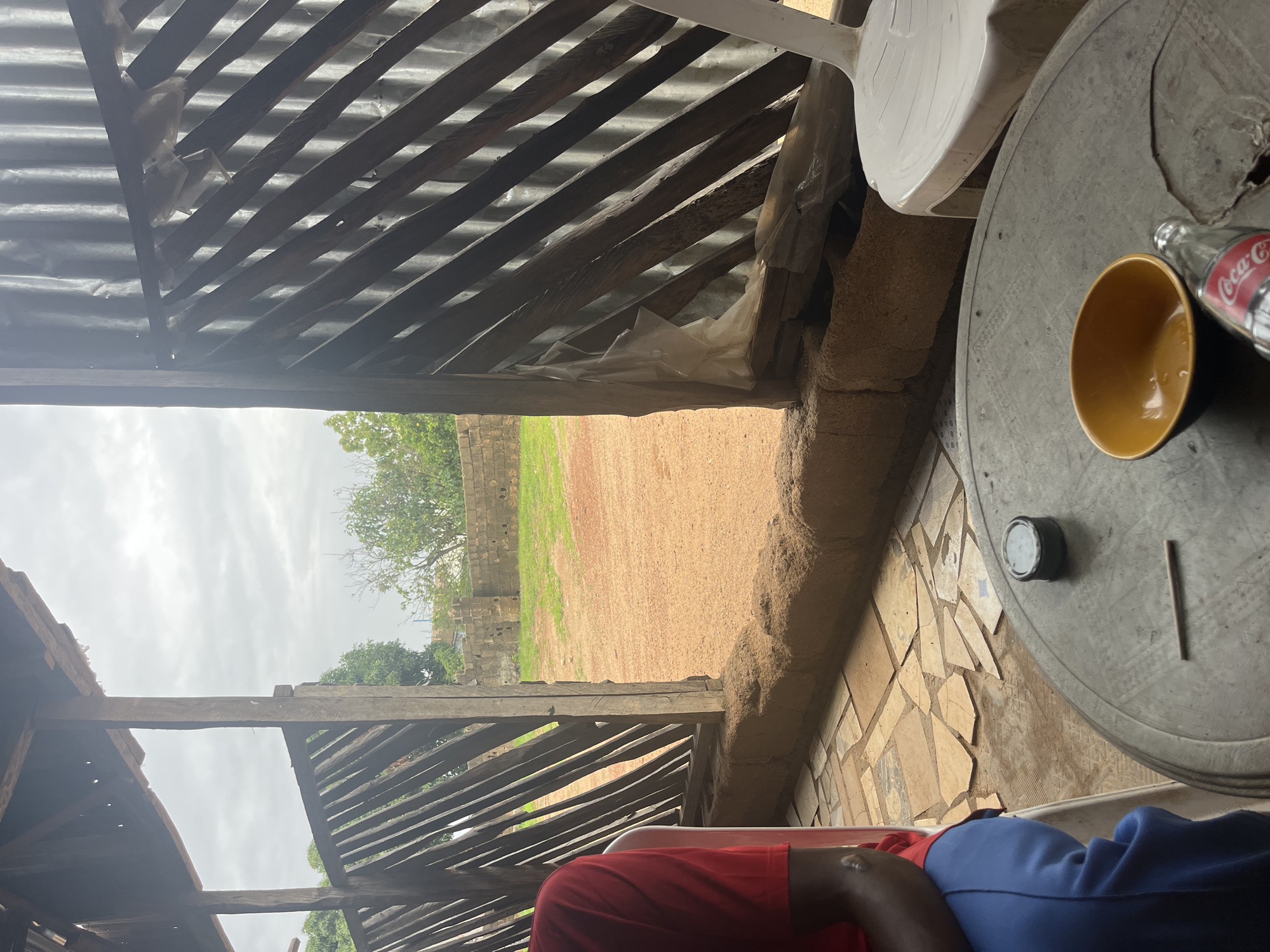 The image captures an outdoor seating area, likely a roadside eatery or a casual gathering spot, situated in the city of Jos, Nigeria. The foreground is dominated by a round, grey table with visible wear and tear. On the table are a small, yellow bowl with residual liquid, a glass bottle of Coca-Cola with its label partially visible, and a single dark-colored stick-like object. To the left of the table, a person is seated, wearing a red t-shirt and blue trousers. They are partially visible from the back and side, with their left arm resting on their leg. A white plastic chair is positioned to the right of the table.
The seating area is sheltered by a rustic structure constructed from wood and possibly corrugated metal on the right side. The walls of the structure are a lattice of wooden planks, creating an open, airy feel. The ground beneath the seating area is paved with irregularly shaped stone tiles. Beyond the structure, an open, sandy area is visible, bordered by a low, rough-textured wall. In the background, a brick wall with visible holes stands amidst sparse green grass and scattered trees. The sky is overcast, suggesting cloudy weather. The overall scene evokes a sense of informal, everyday life in a Nigerian urban or peri-urban environment.