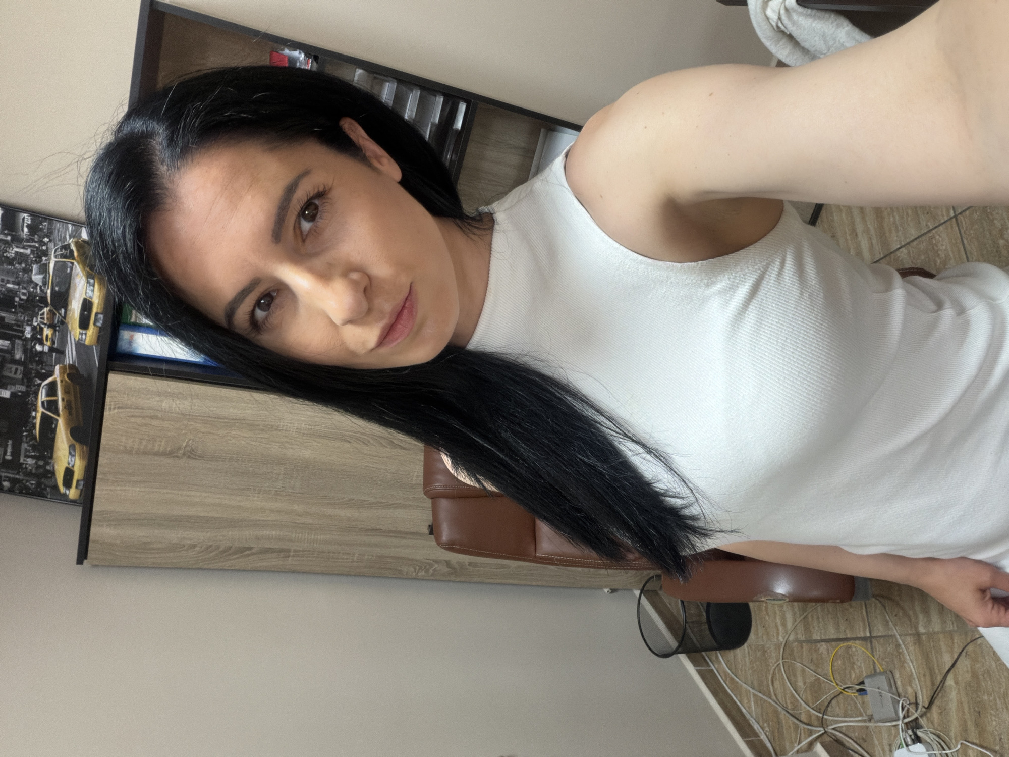 A woman with long, dark hair and fair skin is shown in a selfie. She is wearing a sleeveless white top. Behind her is a light brown wooden cabinet with a black shelf unit above it. A photograph with yellow cabs is visible on the cabinet. A brown leather chair is also partially visible. Wires are scattered on the tiled floor near the chair. The location is Pleven, Bulgaria.
