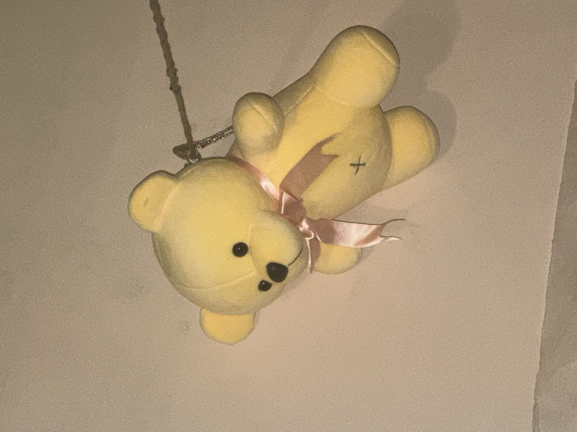 A yellow teddy bear with a pink ribbon is hanging from a string against a textured wall. The teddy bear is facing towards the left side of the image and its left arm is raised. A small black cross is stitched onto its belly. The background is a neutral, sandy color, and the lighting suggests an indoor setting with a light source from the right. There are no people or other objects visible in the scene.
