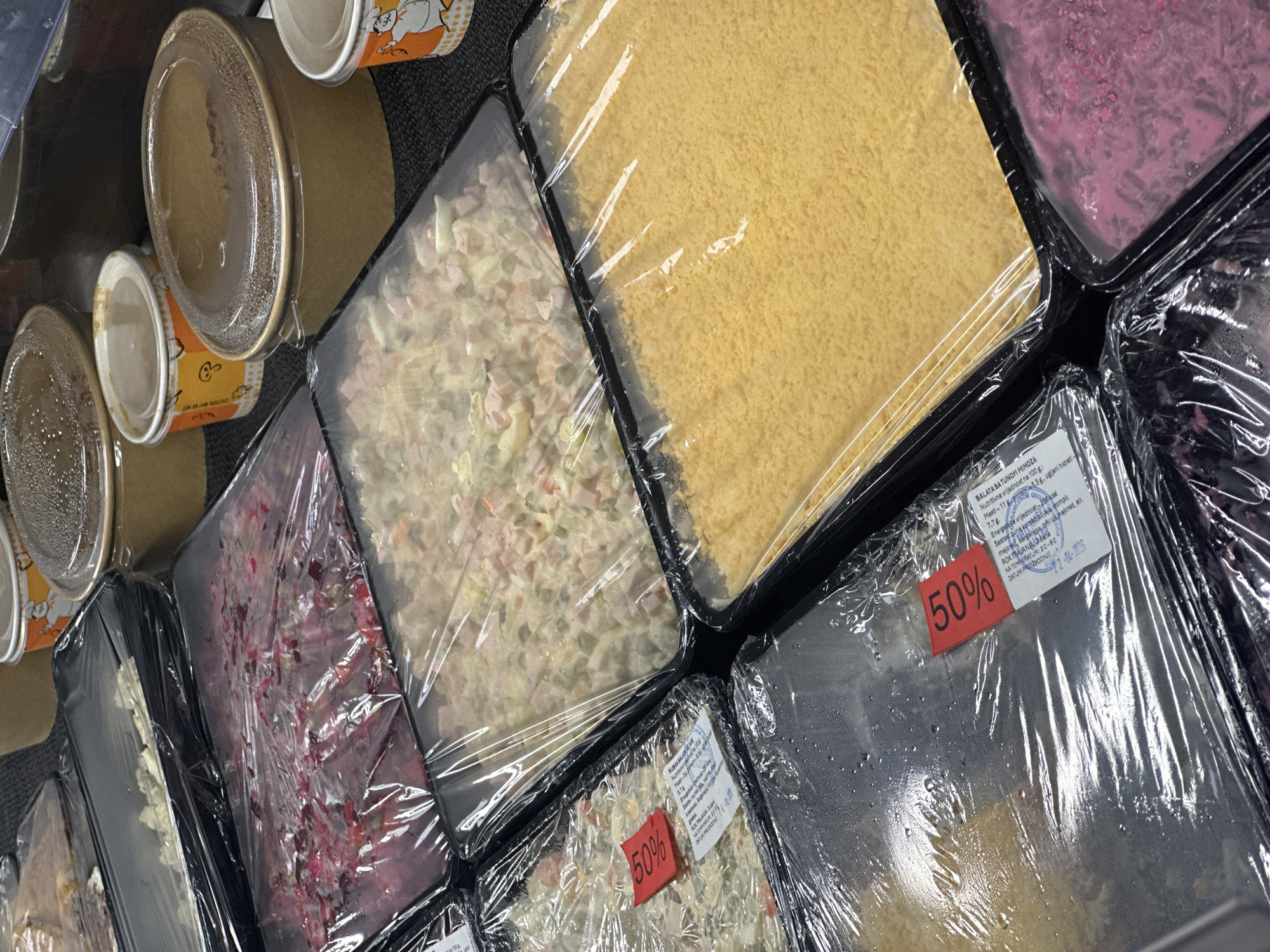 The image captures an overhead view of an assortment of pre-packaged food items, likely displayed for sale in a store in Budva, Montenegro. The scene suggests a deli, supermarket, or take-out counter specializing in ready-to-eat meals and salads.
The subjects are various dishes contained in black plastic trays and a few cardboard cups, all sealed with clear plastic wrap or lids. Condensation is visible under the plastic wrap on many items, indicating refrigeration.
Notable details include:
- Several cardboard cups, one with a prominent "CAUTION MAY BE HOT" warning and a cartoon chicken graphic.
- A large tray filled with a yellow, granular or shredded ingredient, possibly cheese or a breadcrumb topping.
- Trays containing different salads: one appears to be a light-colored, diced salad (resembling an Olivier salad) with ingredients like potatoes and meat, and another is a vibrant pink/purple salad, likely beet-based.
- Two different trays of salad are marked with red "50%" discount stickers, suggesting they are on sale, possibly due to approaching expiry dates.
Visible text on a detailed label belonging to one of the discounted salads provides comprehensive information:
- Product name: "SALATA SA TUNOM MIMOZA" (Tuna Mimosa Salad).
- Nutritional value per 100g: "Masti - 11.8," "prote 5.5g," "ugljeni hidrati - 7.7g," "Energetska vrijednost 236 kcal."
- Ingredients ("Sastav"): "tunjevina komadi, jaja, krompir, majonez, sargarepa, crni luk, majonez, sir" (tuna pieces, eggs, potatoes, mayonnaise, carrots, red onion, mayonnaise, cheese).
- Shelf life: "ROK TRAJANJA 3 dana" (Shelf life 3 days).
- Storage temperature: "NA TEMPERATURI: 2C - 6C."
- Production date: "DATUM PROIZVODNJE 22-04-2026." (Note: The year "2026" appears to be an anomaly given the "3 days" shelf life, possibly a misprint or OCR error for "2024").
Another partial label mentions "RUBKA BALAT" and partial numerical data.