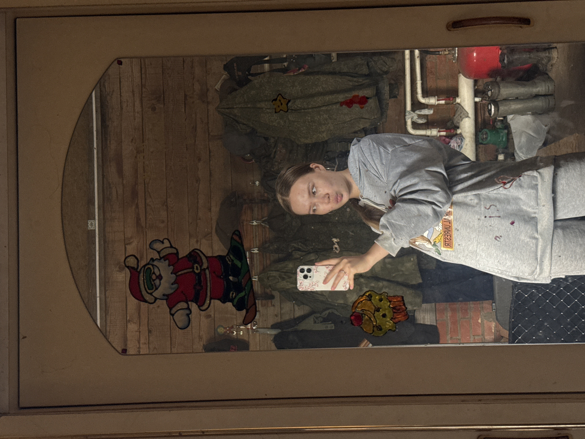 A young woman in Zhigalki, Russia, captures a mirror selfie in what appears to be a utility or storage room. She has light brown hair, pulled back, and is wearing a grey long-sleeved top with mirrored text that reads "ЯГУАРЫ" (Jaguars) and matching grey sweatpants. She holds a white smartphone with a floral-patterned case in her right hand, looking directly at it with a neutral expression.
The reflection in the mirror reveals the interior space. Behind the woman, several pieces of camouflage clothing, possibly military or hunting attire, hang on a wooden plank wall. One jacket features a yellow star patch and a red patch. To the right, exposed plumbing or heating system elements are visible, including a prominent red expansion tank, white pipes, and green valves, set against a background of red brickwork. A black quilted mat can be seen on the floor beneath these fixtures.
The surface of the mirror is decorated with two festive gel clings: a red Santa Claus on the left and a yellow and orange cartoon character, resembling a cookie or treat, on the right. The mirror is encased in a light brown frame, with a small red object on its upper left edge and a wooden handle above it, suggesting it might be part of a cabinet or door. The scene depicts a casual, indoor moment in a functional setting.