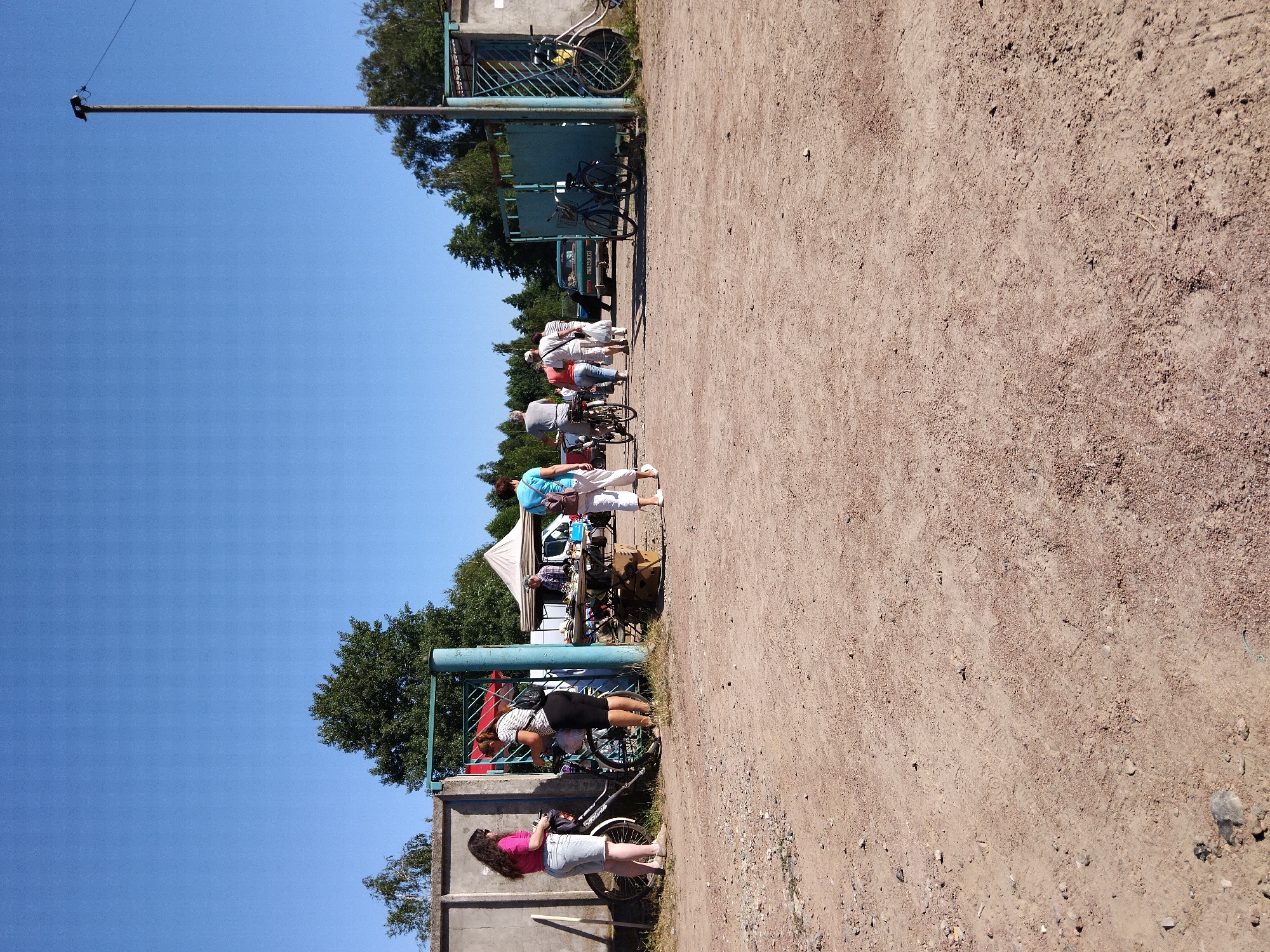 The image shows an outdoor scene with a clear blue sky and a dirt ground. In the foreground, the dirt is scattered with small rocks and debris. In the middle ground, a group of people are present, some of whom are near bicycles. There are also several parked cars and a tent in the background. The scene appears to be a market or gathering, possibly a flea market, given the presence of stalls and people browsing. The environment is rural or semi-rural, with trees visible behind the structures. The overall impression is of a casual daytime event under bright sunlight. It is possible that this scene is located in the city of Irshansk, Ukraine, as indicated in the prompt, however, there are no specific visual cues within the image to confirm this location.