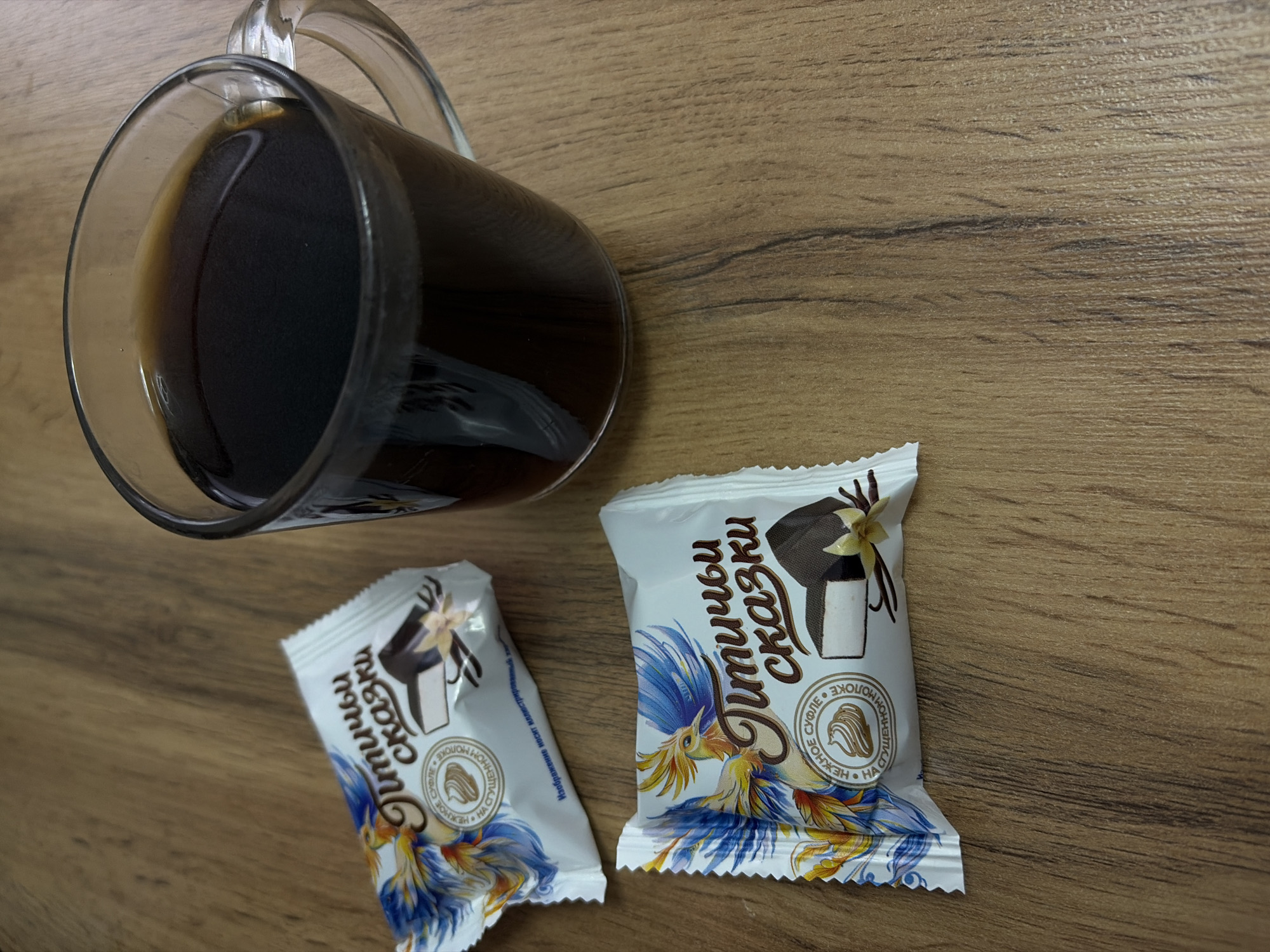 The image shows a wooden table surface with a glass mug containing a dark liquid, likely coffee or tea, and two individually wrapped candies labeled "Птичьи сказки" (Ptichye Skazki, "Bird's Tales"). The candies are white with images of a dessert and a blue and yellow bird on the packaging. The candies also include the texts 'НЕЖНОЕ СУФЛЕ' and 'СУЩЕННОМ МОЛОКE', indicating they are soufflé candies made with condensed milk.
Based on the text on the packaging, the candies are a product of the Chaikovsky Confectionary. The environment suggests an indoor setting, possibly a home or office. There are no people present in the frame. The interaction is implied through the arrangement of the objects, suggesting a coffee break or snack setting.