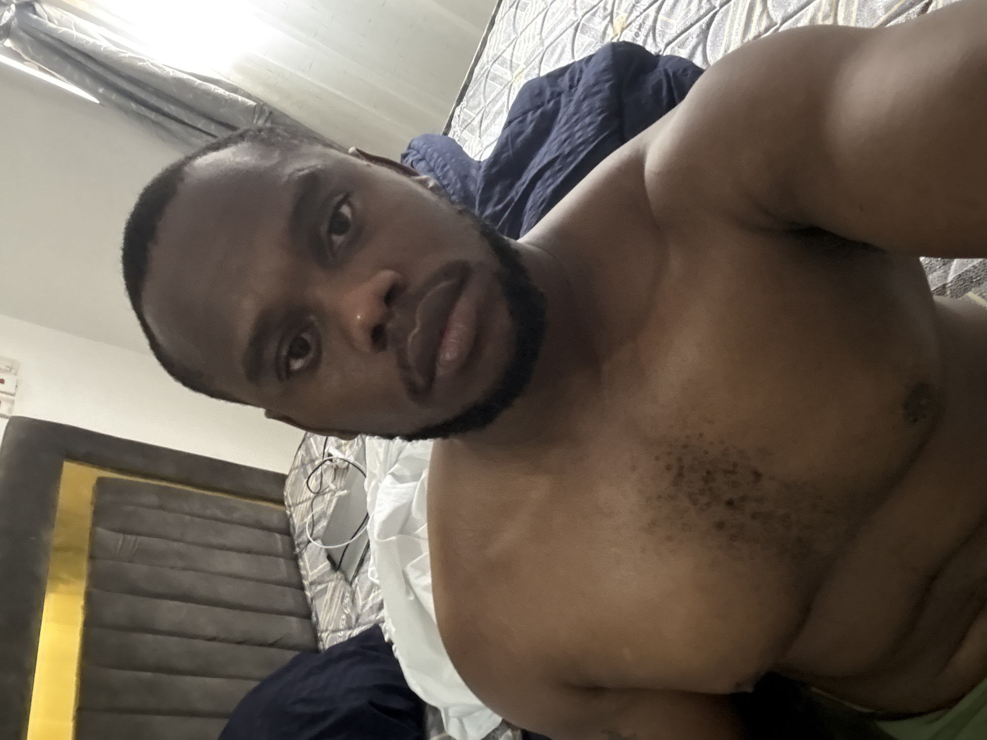 A shirtless man with dark skin and a beard is reclining on a bed. The bed has a dark gray headboard with gold trim and a patterned gray and white comforter. A blue blanket is also on the bed. A white object with cords (possibly a fan or a lamp) is visible behind the man. The setting appears to be indoors, possibly a bedroom. The exact location within the city of Iddo Tudunwada, Nigeria cannot be determined from the given image.