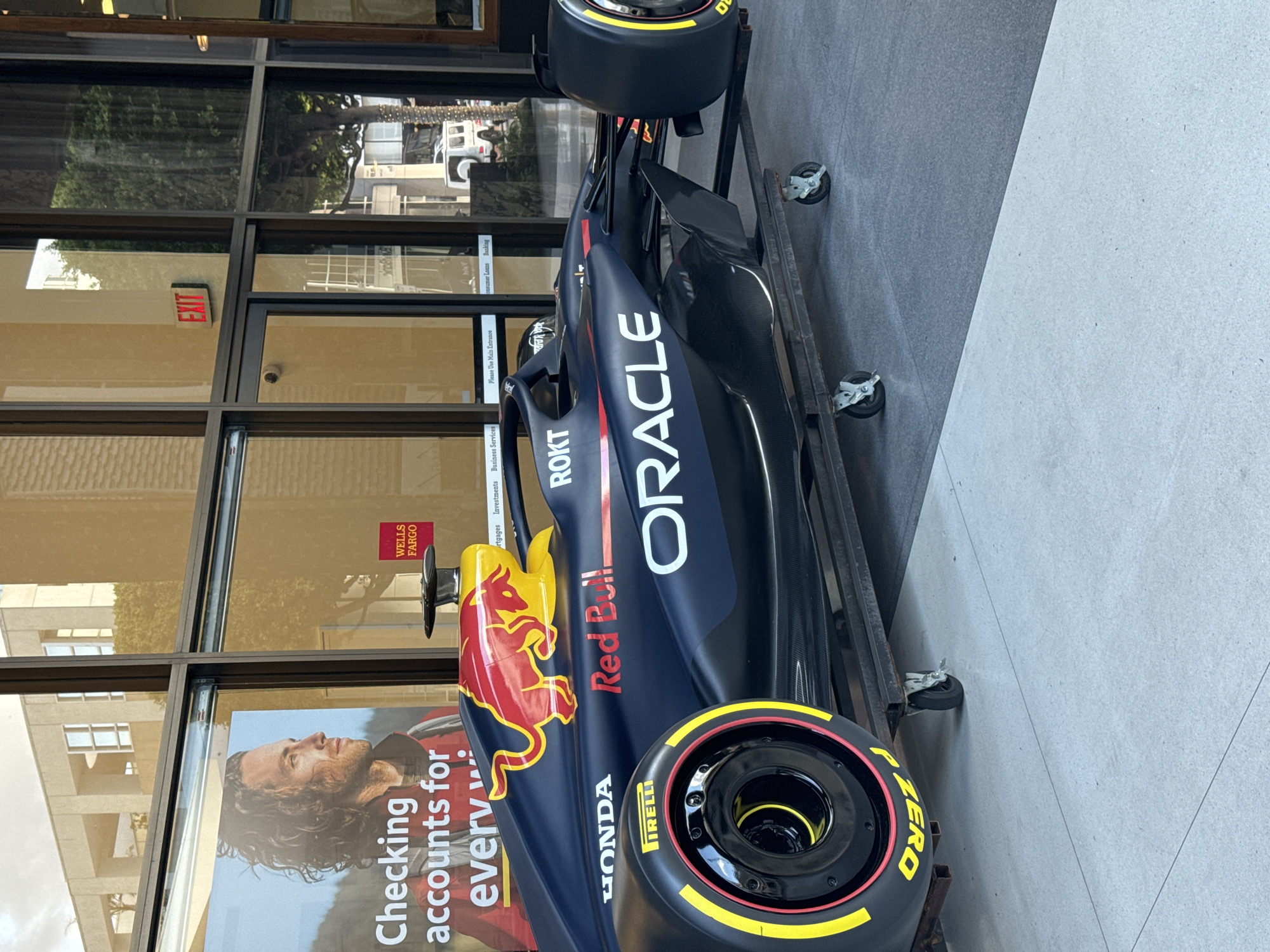 The image captures a Red Bull Racing Formula 1 car displayed in front of a Wells Fargo bank branch in Beverly Hills, California. The car is positioned on a mobile platform with wheels, allowing it to be moved. The livery features the prominent "ORACLE" branding, along with "Red Bull" and "HONDA" logos. The car's tires are Pirelli "Zero". The bank's window display includes an advertisement promoting checking accounts. The scene is captured during daylight hours, evidenced by the natural lighting and reflections in the bank's windows.