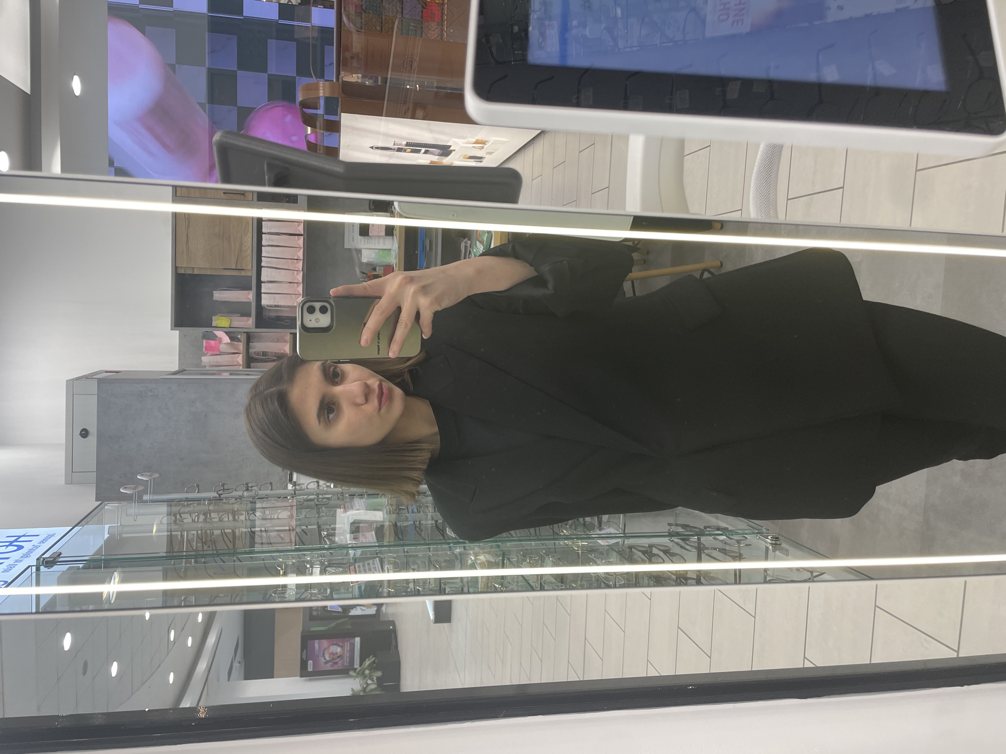 A woman with brown hair and light skin, dressed in a black blazer, holds a smartphone and takes a selfie in front of a store display case for eyeglasses. The store seems to be brightly lit with white tiled floors and ceiling lights. The location appears to be an eyeglasses store in a modern building, and, based on the background and architectural style, the scene is likely in a city like Sofia, Bulgaria.