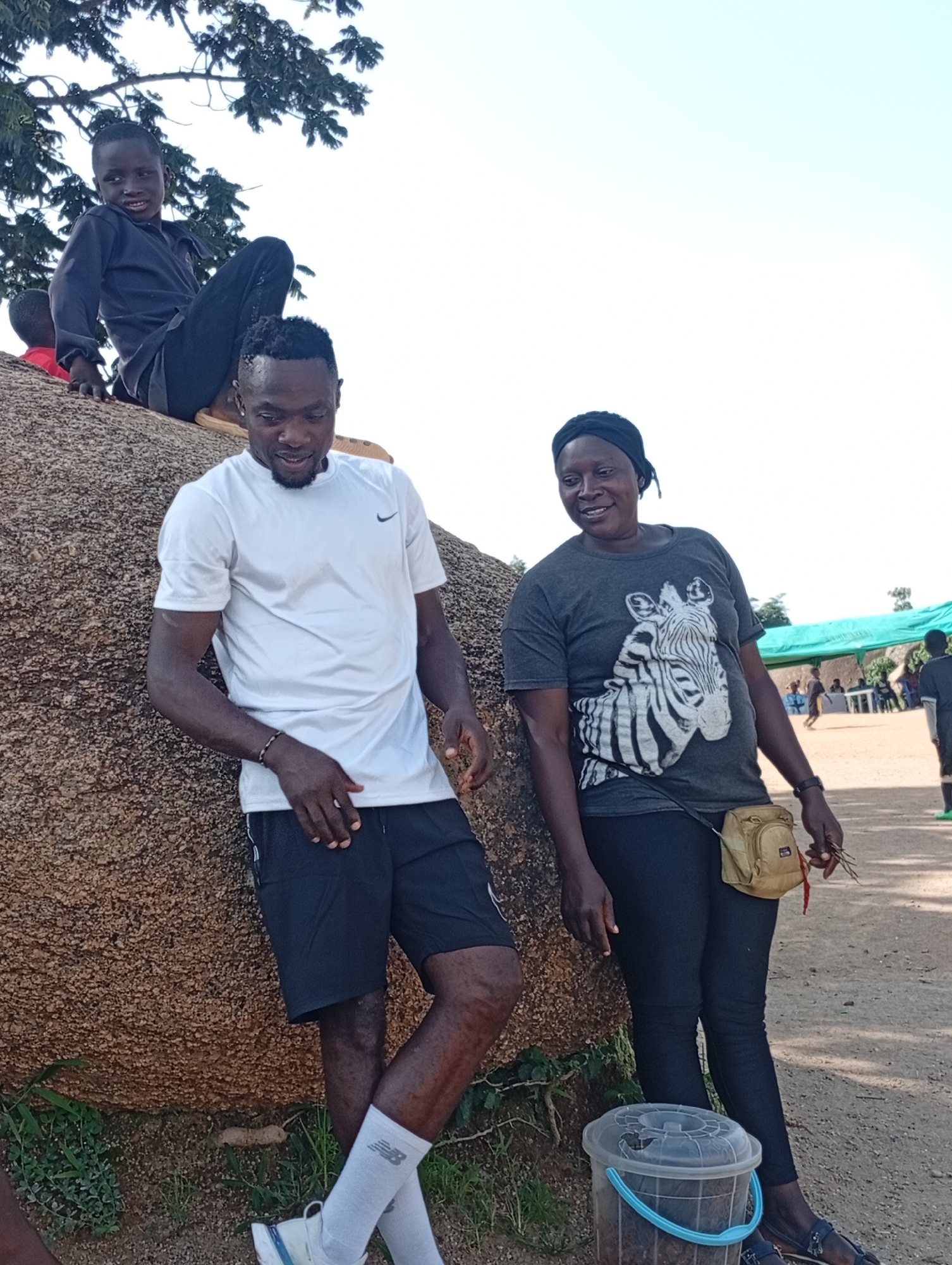 In Unknown, Unknown, two adults and a child are seen on a large, rough rock. A young child in a dark blue long-sleeved shirt and dark pants is sitting on the rock, looking towards the viewer. A man in a white t-shirt and black shorts, wearing white socks and sneakers, leans against the rock. A woman in a dark t-shirt with a zebra print and dark leggings, wearing a black head covering and sandals, stands next to the man, also leaning slightly against the rock. She is holding a small bunch of twigs in her right hand. A tan fanny pack is clipped around her waist. In front of the woman, on the ground, is a clear plastic bucket with a blue handle, filled with dark material. In the background, there is a green tent canopy, with people milling about and a white table. The ground is dirt and pebbles. The sky is bright and clear.