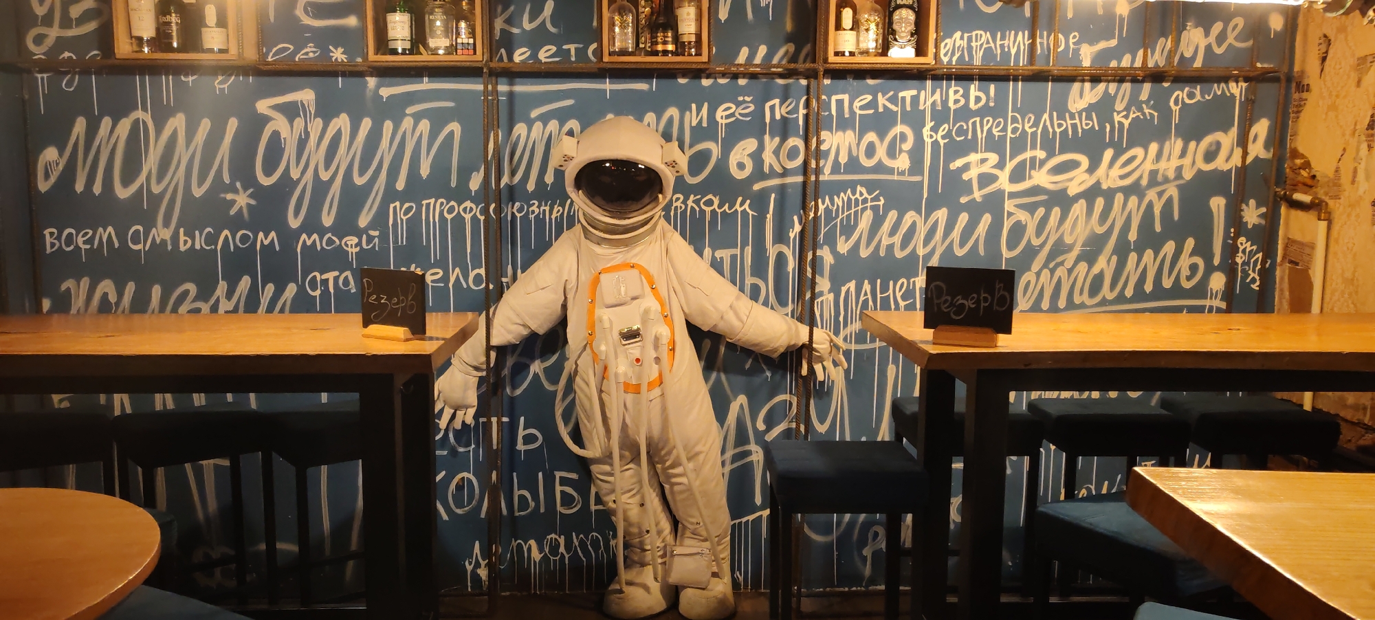 A full-body mannequin dressed as an astronaut stands to the left of center in front of a blue wall with white graffiti. The astronaut is wearing a white suit with orange accents and a helmet with a reflective visor. The wall is covered in various phrases and designs in white spray paint. To the left of the astronaut, a wooden table is set with a small black chalkboard sign that reads "Резерв" (Reserve). Behind the table, several dark blue bar stools are visible. To the right of the astronaut, another wooden table is partially visible, also with dark blue bar stools beneath it. The lighting is dim, with some of the wall and objects illuminated by spotlights. The scene appears to be in a bar or restaurant, possibly in Kaluga, Russia, given the Cyrillic script on the chalkboard.