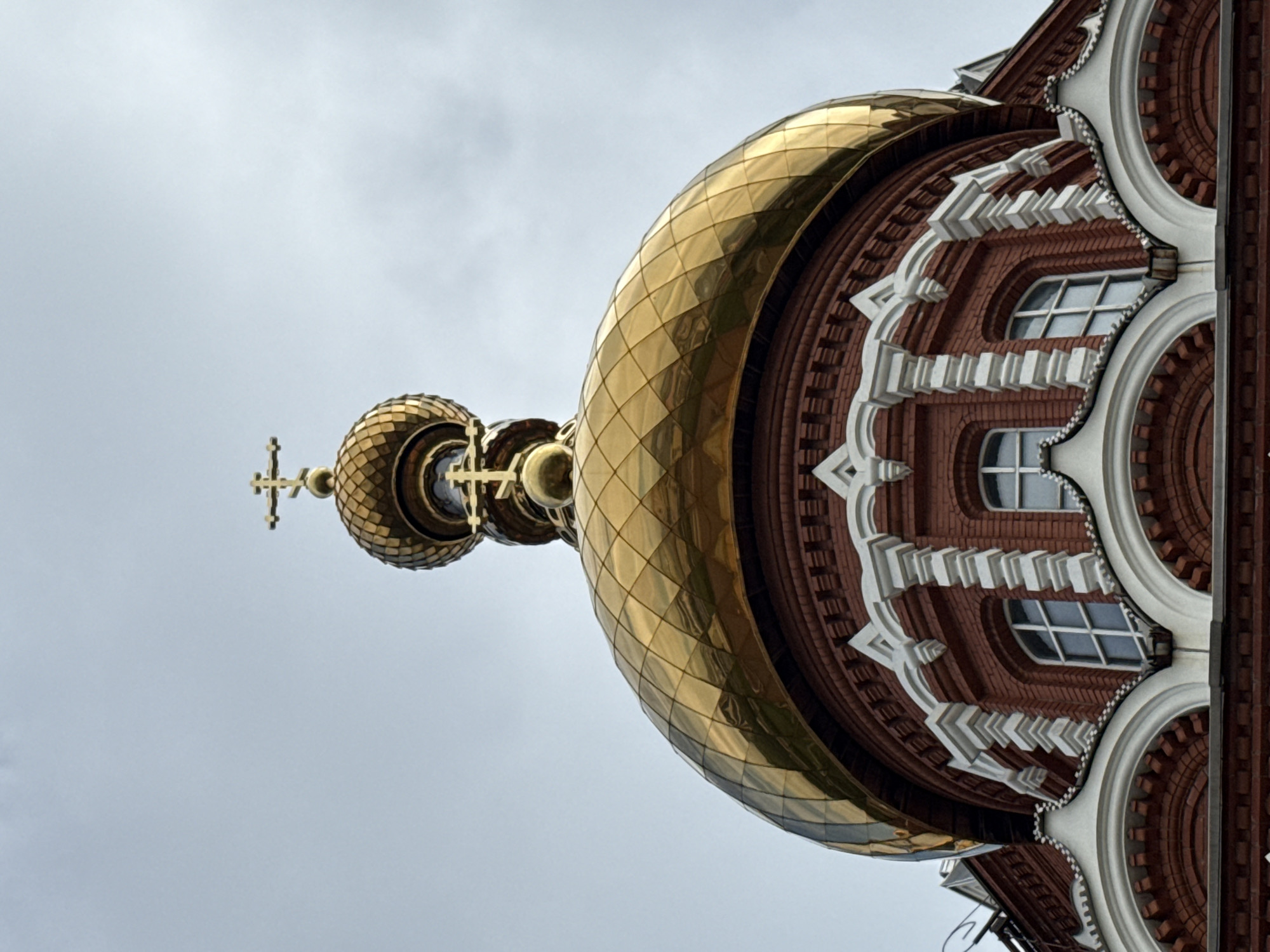 The image shows the golden dome and upper section of the Holy Trinity Cathedral in Izhevsk, Russia. The dome is ornate, with a cross at its apex. The cathedral's facade is constructed of red brick with white decorative elements, including arched windows. The sky is overcast. There are no people visible in the image.