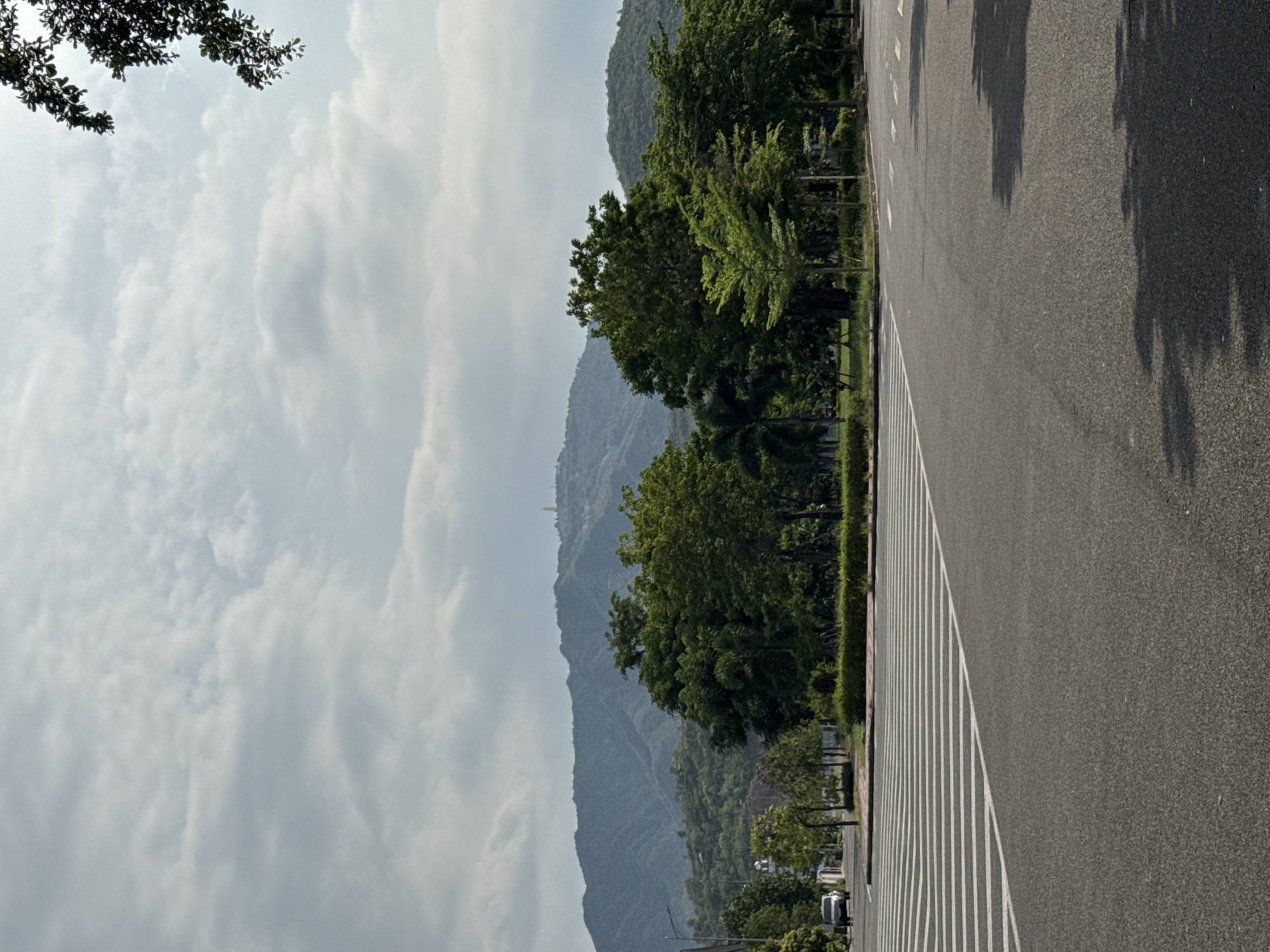 The image depicts a road with a pedestrian crossing in the foreground, leading towards a line of trees and a mountainous backdrop. The sky is overcast with scattered clouds. To the left, a white car is visible on the road, approaching the pedestrian crossing. Further back, there are more trees and some low-rise buildings. The landscape appears to be part of a developed area with green spaces and a prominent mountain range in the distance. The scene suggests a serene environment, possibly on the outskirts of a city or in a suburban area.
