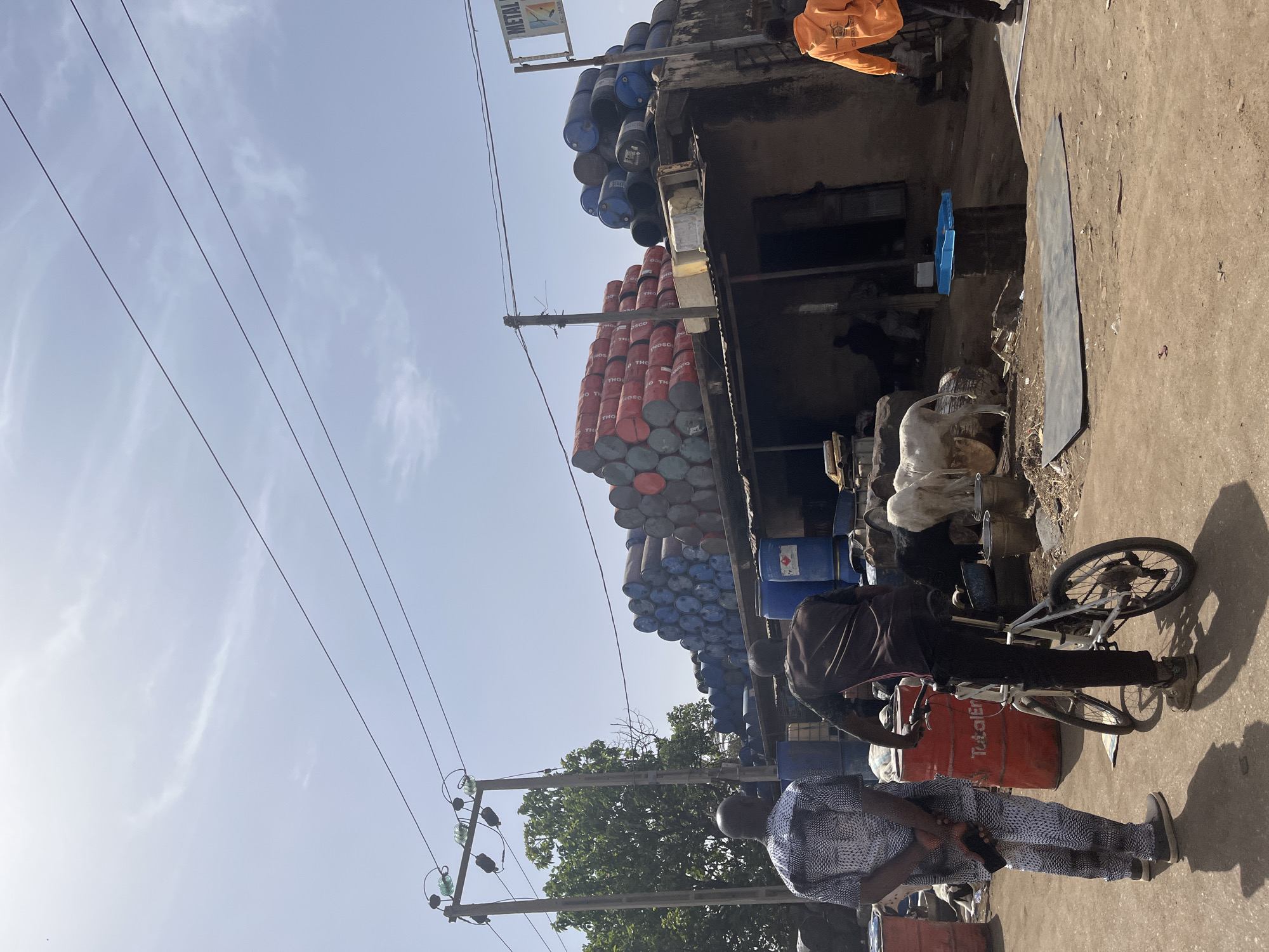 The image captures a scene in Jos, Nigeria, characterized by a roadside commercial establishment. Piles of blue and red metal drums are stacked high against a building. Electrical power lines run parallel above the street. Two men are in the foreground. One man is standing in a patterned shirt and holding a phone. The other man is on a bicycle next to a red drum marked "TotalEn". In the background are various metal objects and containers. The environment appears to be urban, with concrete ground and structures.