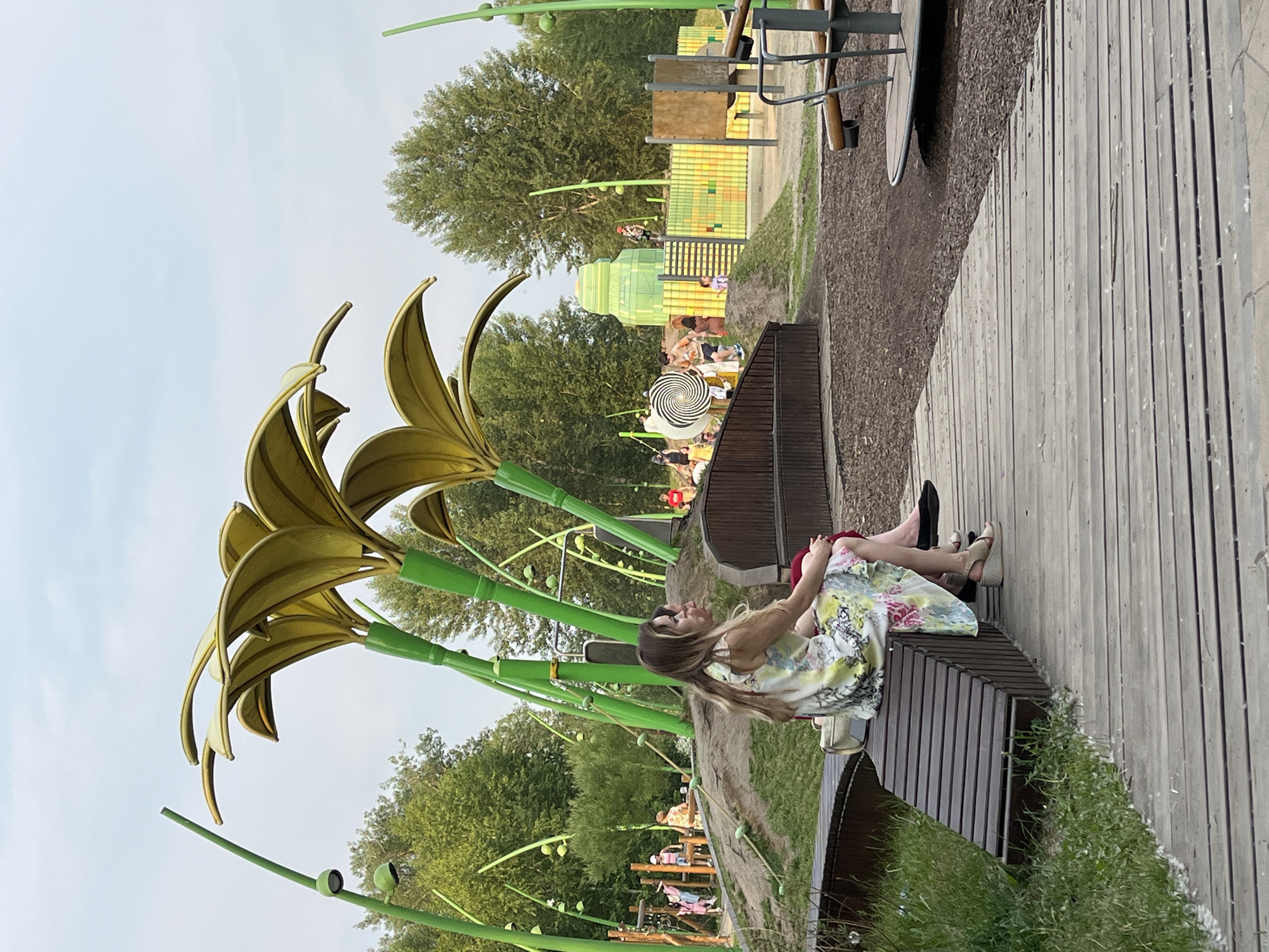 Two women are sitting on a wooden bench in a park. The woman in the foreground is wearing a floral print dress and sandals, and the woman next to her is wearing a red garment. They are both facing to the left, looking at a playground with slides and climbing structures. The playground is decorated with large, yellow, flower-shaped canopies supported by green poles. In the background, there are trees and a cloudy sky. This scene is located in the city of Krasnoyarsk, Russia.