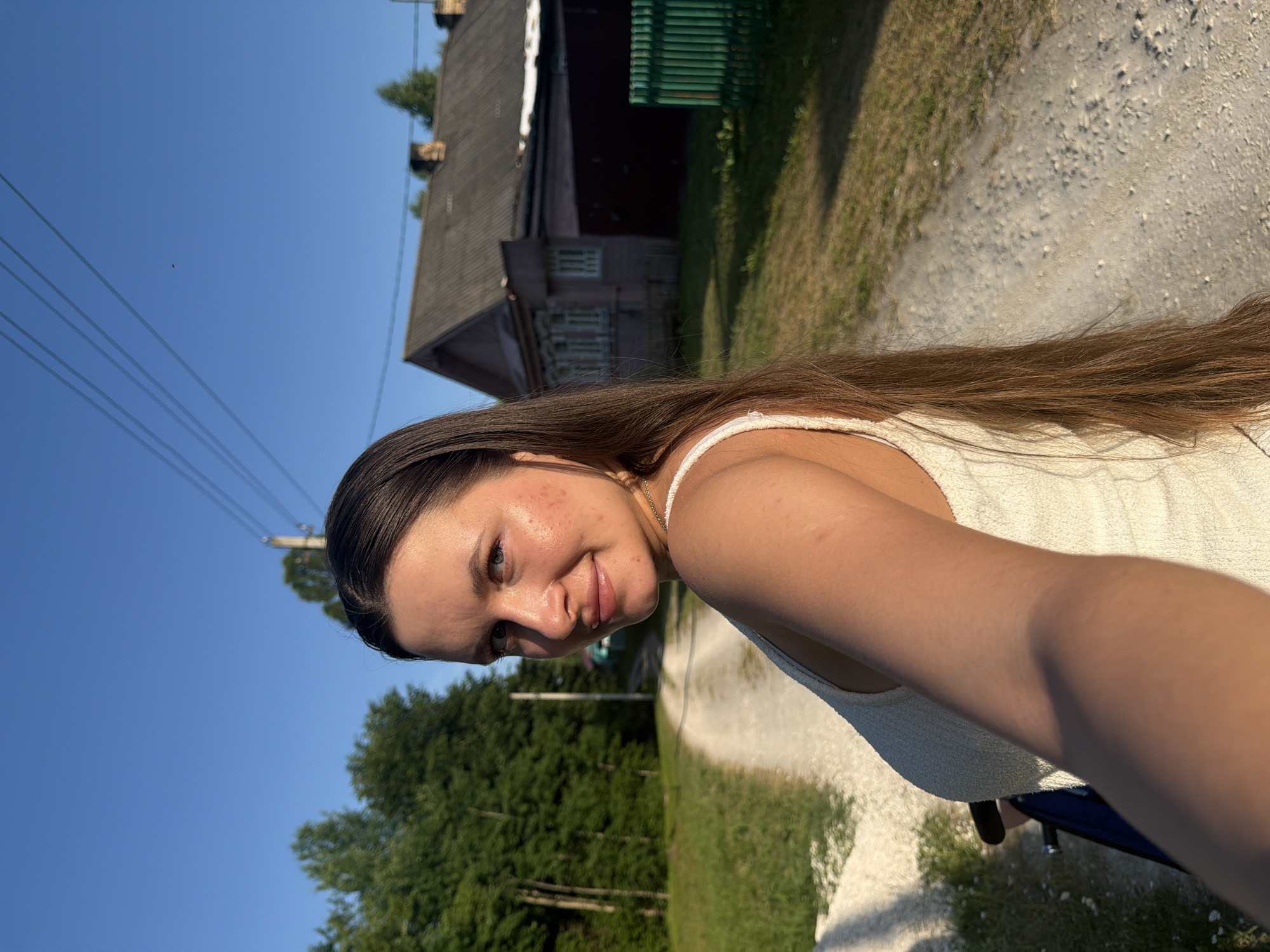 An outdoor, eye-level, medium shot captures a woman with long, dark brown hair and fair skin looking towards the camera while holding a phone in her right hand, likely taking a selfie. She is wearing a white, textured tank top. Her face is turned slightly to the left, revealing blue eyes and a gentle smile. She has freckles across her nose and cheeks, and some minor acne.
The background depicts a rural setting. To the left, a dirt road curves into the distance, flanked by lush green trees and grass. A single utility pole stands near the road. To the right, a weathered, pink wooden house with a dark grey roof is visible, along with a dark brown fence and a bright green fence. The sky above is a clear, bright blue, with a few thin power lines crossing the upper portion of the frame. The lighting suggests it is late afternoon or early evening, with the sun casting warm tones on the scene. The location is identified as Krasny Mayak, Russia.