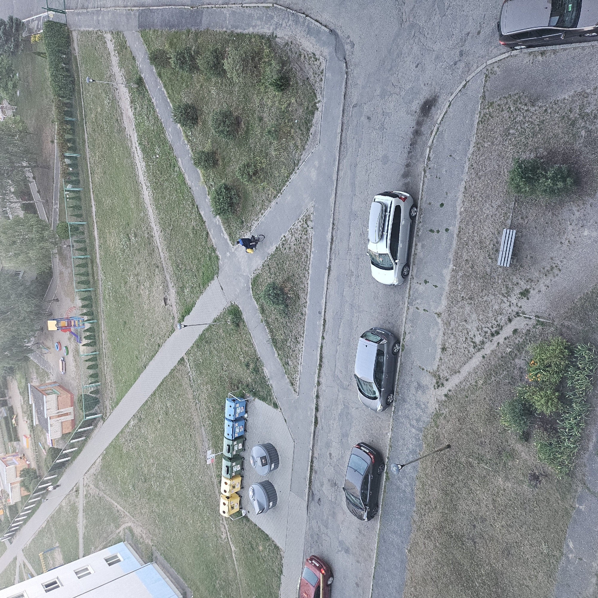 An aerial view captures a residential area in Brest, Belarus, with a multi-story apartment building on the left. A paved pathway winds through a grassy area dotted with trees and bushes. Several cars are parked along the roadside, including a red sedan, a dark sedan, a gray hatchback, and a silver station wagon with a roof rack. Across from the apartment building, a row of recycling bins is visible, along with two larger cylindrical waste containers. In the distance, a playground with a slide and swings is situated near another building. A person on a bicycle with a yellow bag on the back is riding along the pathway. A park bench is visible in the grassy area to the right.