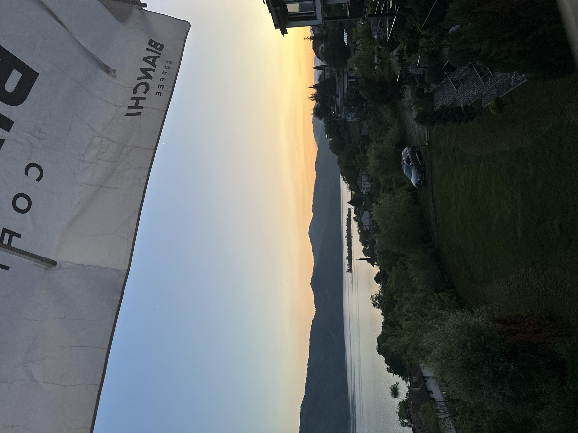 The image presents an elevated, panoramic view of Glavatartsi, Bulgaria, at twilight, overlooking a large body of water and mountainous terrain. The upper portion of the frame is dominated by the underside of a white fabric umbrella, upon which mirrored black text is visible. When un-mirrored, the text reads "BIANCHI COFFEE."
Below the umbrella, the sky displays a clear gradient, transitioning from a light blue at the zenith to a warm orange and yellow hue along the horizon, indicative of sunset or sunrise. A long, dark silhouette of mountains forms the distant backdrop, bordering the calm, reflective surface of the water, which appears to be a lake or reservoir.
Along the immediate shoreline and hillside, a residential area of Glavatartsi is visible. Houses with light-colored facades and dark roofs are interspersed among dense clusters of green trees and vegetation. A prominent, slender dark structure, likely a minaret, stands out against the water on the right side of the lake. To the left of the minaret, a small grouping of dark, possibly submerged, trees or structures is visible in the water.
In the lower right foreground, part of a multi-story building with dark balconies and windows is visible. Adjacent to this structure, a winding cobblestone path descends, flanked by manicured shrubs and trees. To the left of the path, a well-maintained green lawn slopes downwards. A dark-colored SUV is parked on a dirt patch at the edge of the lawn. The overall lighting in the lower portion of the scene is dim, consistent with early evening. No people are visible in the image.