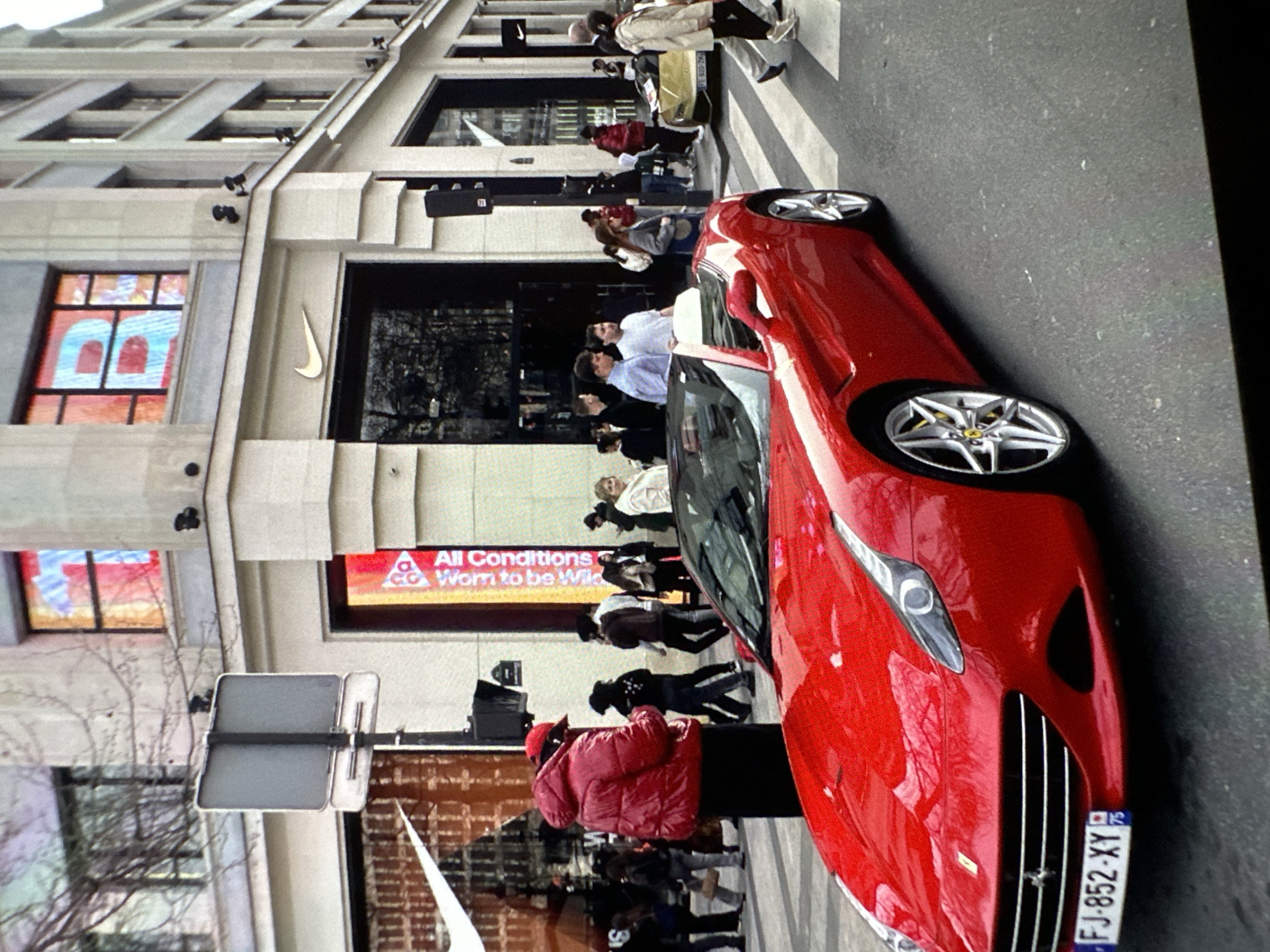 The image shows a red Ferrari parked on a street in front of a Nike store. Pedestrians are walking on the sidewalk and crossing the street. The street is paved with asphalt. The building is multi-story, with windows and architectural details. A street sign is visible. The license plate of the car reads "FJ-852-XY." The location is likely not Chișinău, Moldova as the license plate appears to be French.