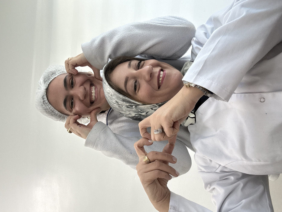 The image displays two women in an indoor setting in Meknes, Morocco. Both individuals are smiling broadly. The woman in the foreground wears a white lab coat with visible metal snap buttons, a grey and white patterned headscarf, a silver-colored watch on her left wrist, and multiple rings on her left hand, including a gold-colored ring on her right index finger. Her hands are clasped together, with fingers curved to form the lower portion of a heart shape. The woman positioned behind and slightly above her wears a white disposable hairnet and a light grey long-sleeved top. Her hands are positioned near her cheeks, with thumbs and index fingers forming small heart shapes on both sides of her face. She wears gold-colored rings on her index fingers and gold-colored hoop earrings. The background consists of a plain, light-colored wall, indicative of a professional or controlled environment.