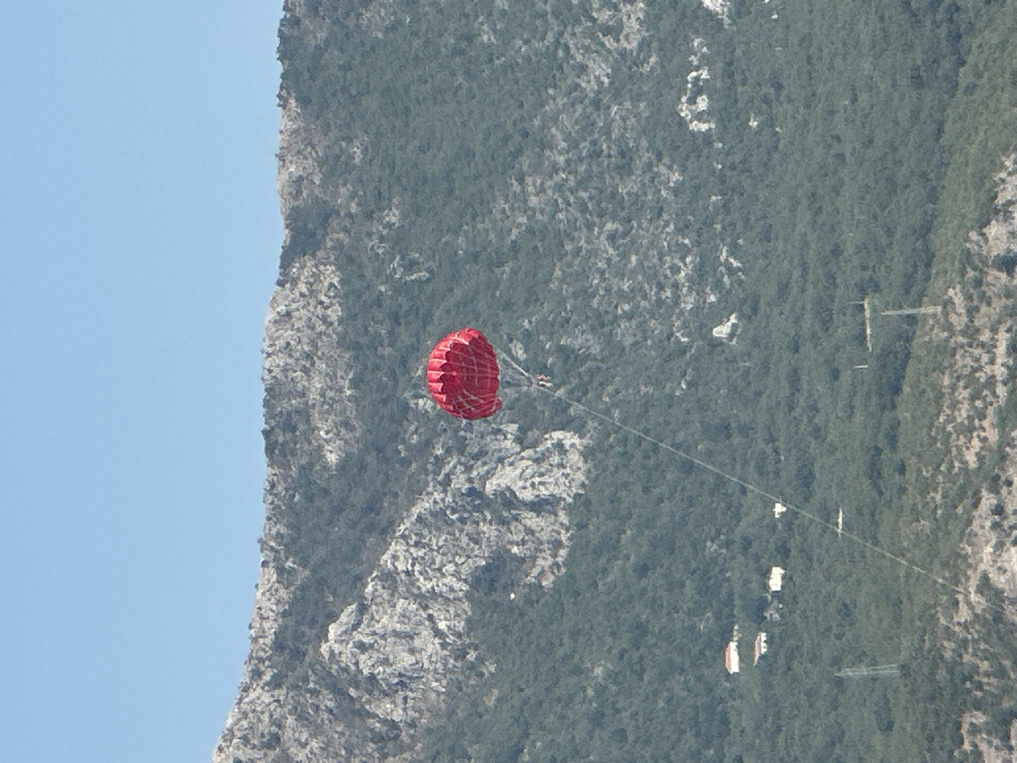 The image shows a red parachute with a person underneath it in mid-air. The parachute is against a backdrop of a tree-covered mountain and a clear blue sky. In the distance, several white buildings with red roofs are visible on the mountainside. There are also power lines in the lower part of the image. The overall scene suggests a recreational activity taking place in a mountainous coastal area, likely near Boreti, Montenegro, given the landscape.
