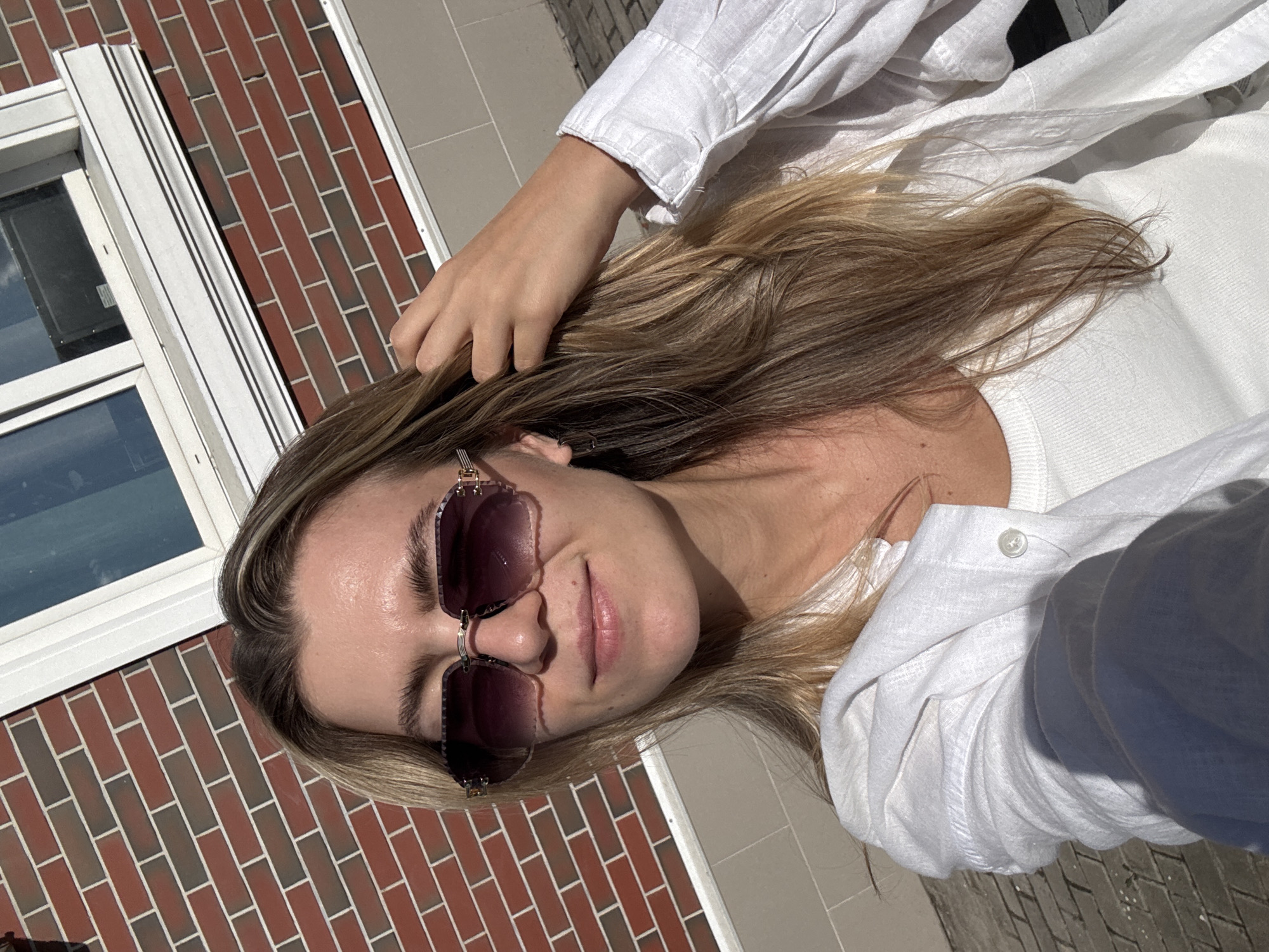 An outdoor, close-up, selfie-style photograph shows a light-skinned young woman with blonde hair, wearing oversized, dark-tinted sunglasses and a white button-down shirt over a white tank top. Her left hand is raised and touching her hair. She is standing outdoors with a red brick building and a window in the background. The ground appears to be paved with interlocking pavers. The lighting suggests it is daytime, and the sun is casting shadows.