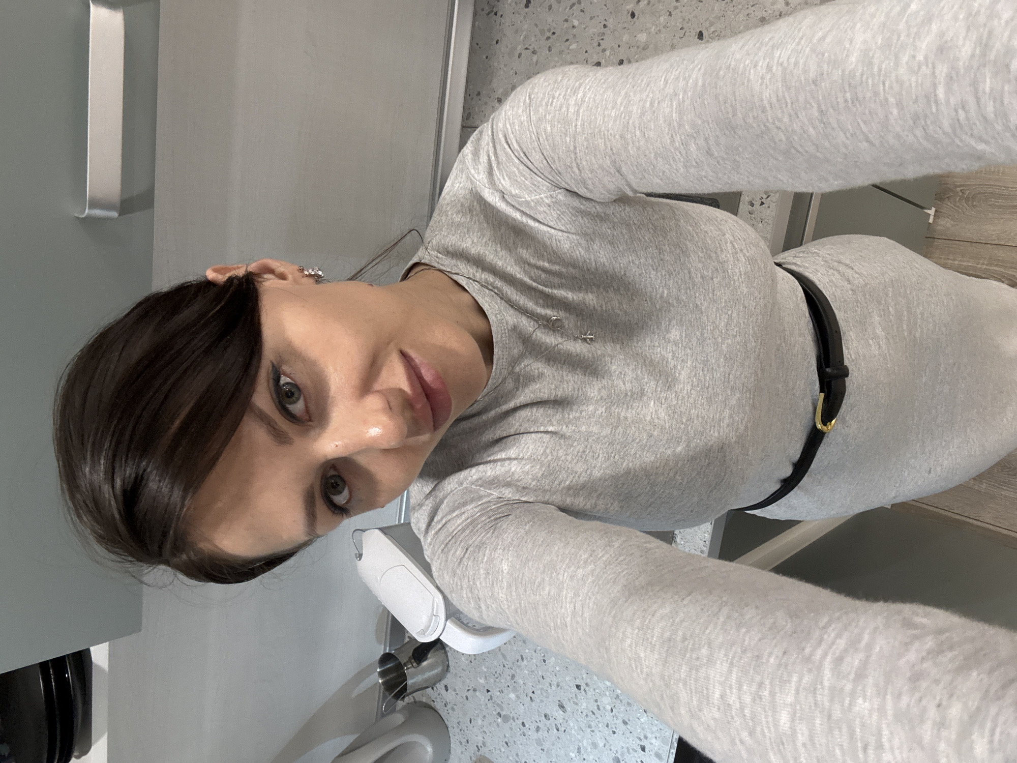 A woman is in a room with light gray walls and countertops with speckled gray and white patterns, possibly a kitchen or bathroom. She is wearing a long-sleeved, light gray dress with a black belt. A stainless steel kettle and a white electric kettle are on the countertop. The location is potentially Murino, Russia.