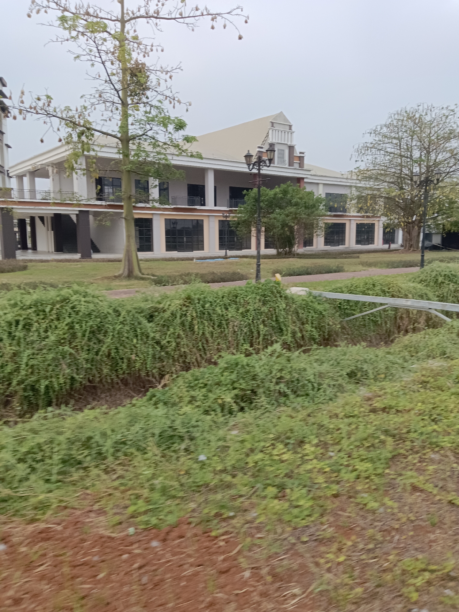 A long, multi-story building with a white facade and a cream-colored roof is visible in the background. The building has a symmetrical design with large windows and balconies. In the foreground, there is a lush green landscape with various trees and bushes. A brown dirt path runs through the scene, and there is a metal railing in the middle ground. The sky is overcast, suggesting a cloudy day. The image appears to be taken in the city of Jayamukti, Indonesia.