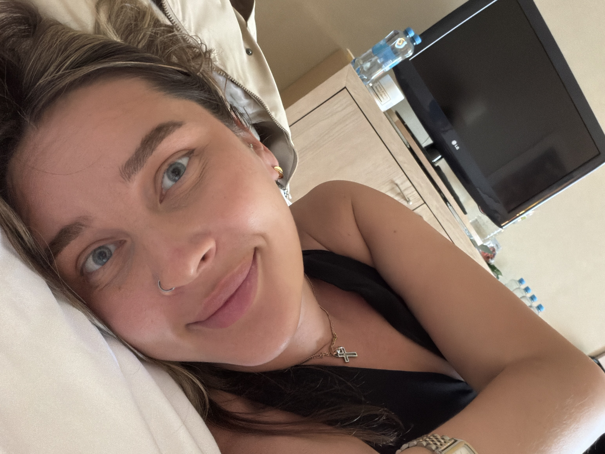 A person is lying down, possibly in a bed, with a white pillow visible under their head. They have light-colored eyes, a nose ring, and are wearing a necklace with a cross pendant. The person's hair is light brown with darker roots. They are wearing a dark-colored top. In the background, there's a television set mounted on a wall or a piece of furniture, a cabinet with water bottles on top. The scene doesn't contain information to be specifically located in Moscow, Russia.