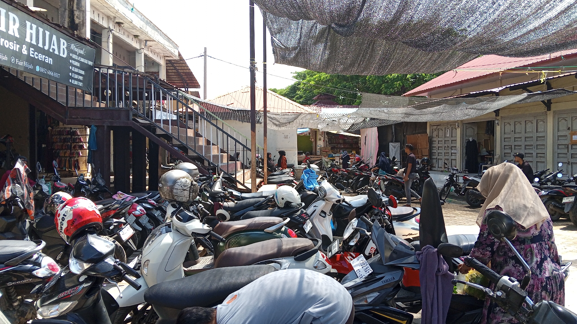 The image depicts a busy marketplace or street scene in Lhokseumawe, Indonesia, likely an area with shops and parking for motorcycles. In the foreground, a dense collection of parked motorcycles dominates the lower half of the frame. Several individuals are present, some appearing to be shoppers or vendors.
On the left, a building with a prominent sign reading "FAIR HIJAB" is visible, with stairs leading up to an upper level. This building appears to be a shop selling hijabs and other clothing items, as indicated by racks of folded goods displayed behind a glass pane on the ground floor.
In the center and right of the image, more buildings with storefronts are lined up, some with awnings or protective netting overhead. The scene suggests a commercial area with high pedestrian and vehicle traffic, typical of a bustling Indonesian city. The atmosphere is sunny, with shadows cast by the overhead coverings and buildings.