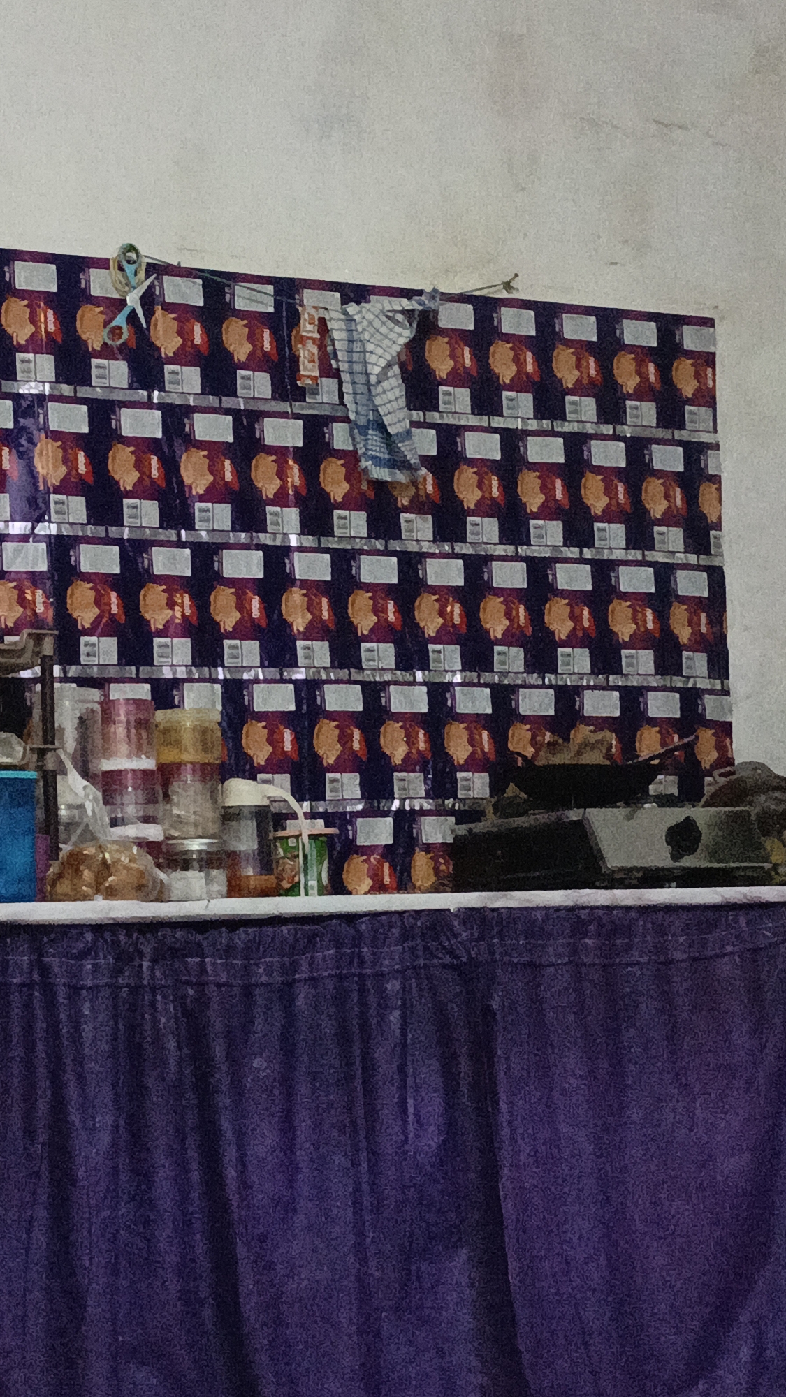 The image shows a shelf of snack packages mounted on a wall. The packages are all the same, featuring a purple background with an orange circular design and white text. To the left of the packages, a pair of blue scissors with a green rubber band is hanging from a wire. A blue and white checkered cloth is also hanging from the wire to the right of the scissors. Below the shelf, a purple table cloth covers a surface, on which several containers and a stove are visible. The stove has a pan on top. The environment appears to be an indoor setting, likely a market stall or a small shop. The image is taken from a slightly low angle, looking up towards the wall.