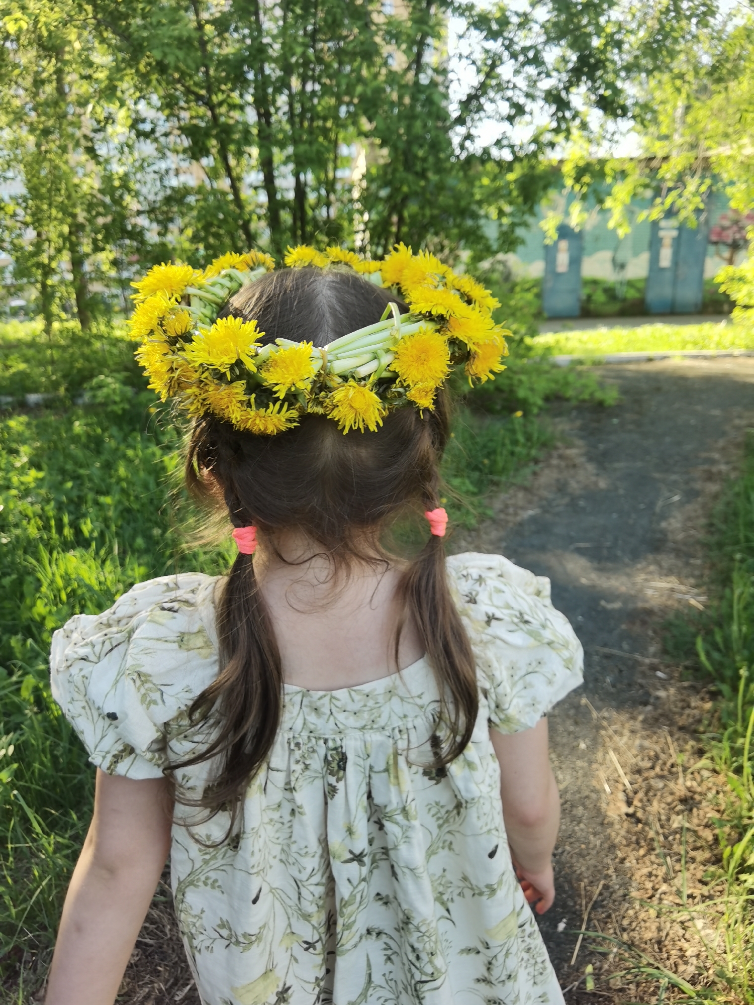 A young girl, viewed from the back, stands on a path in Perm, Russia. She wears a white dress with a green and yellow floral pattern and has brown hair styled in pigtails secured with orange hair ties. A dandelion wreath adorns her head. The path is bordered by grass, and in the background are trees and a building, indicating an outdoor, potentially urban, setting.