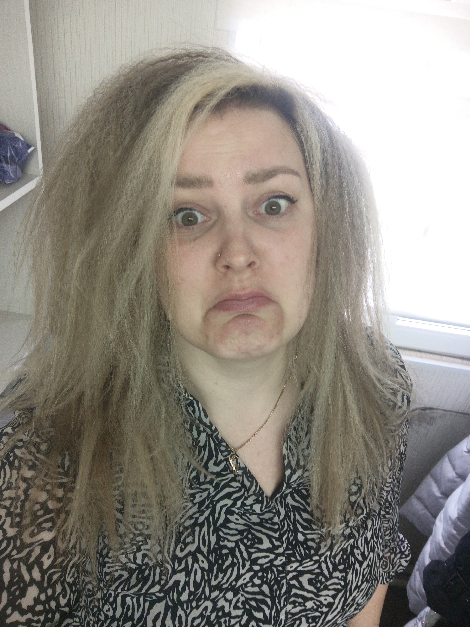 A woman with light blonde crimped hair is shown in a close-up, front-facing selfie. Her expression is one of exaggerated displeasure, with wide eyes and a downturned mouth. She wears a black and white animal print blouse and a thin gold chain necklace. The background suggests an indoor setting with a partially visible white shelf unit on the left and a window on the right. The location is not verifiable as being Noviy, Russia, or any specific location.