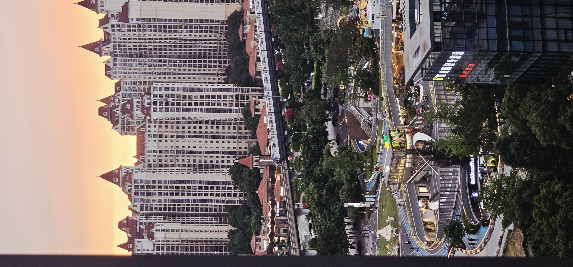 Captured from an elevated perspective, the image presents a view of Liangjiang, China. The scene is divided into two distinct halves: on the left, multiple high-rise apartment buildings stand tall, characterized by their red-tiled roofs and white facades. To the right, a vibrant urban landscape unfolds with walkways, trees, and modern commercial structures. A monorail train is visible, traversing between these two areas. People are seen walking along the paths of the urban area. The light suggests either dawn or dusk, with a soft glow in the sky behind the apartment buildings.