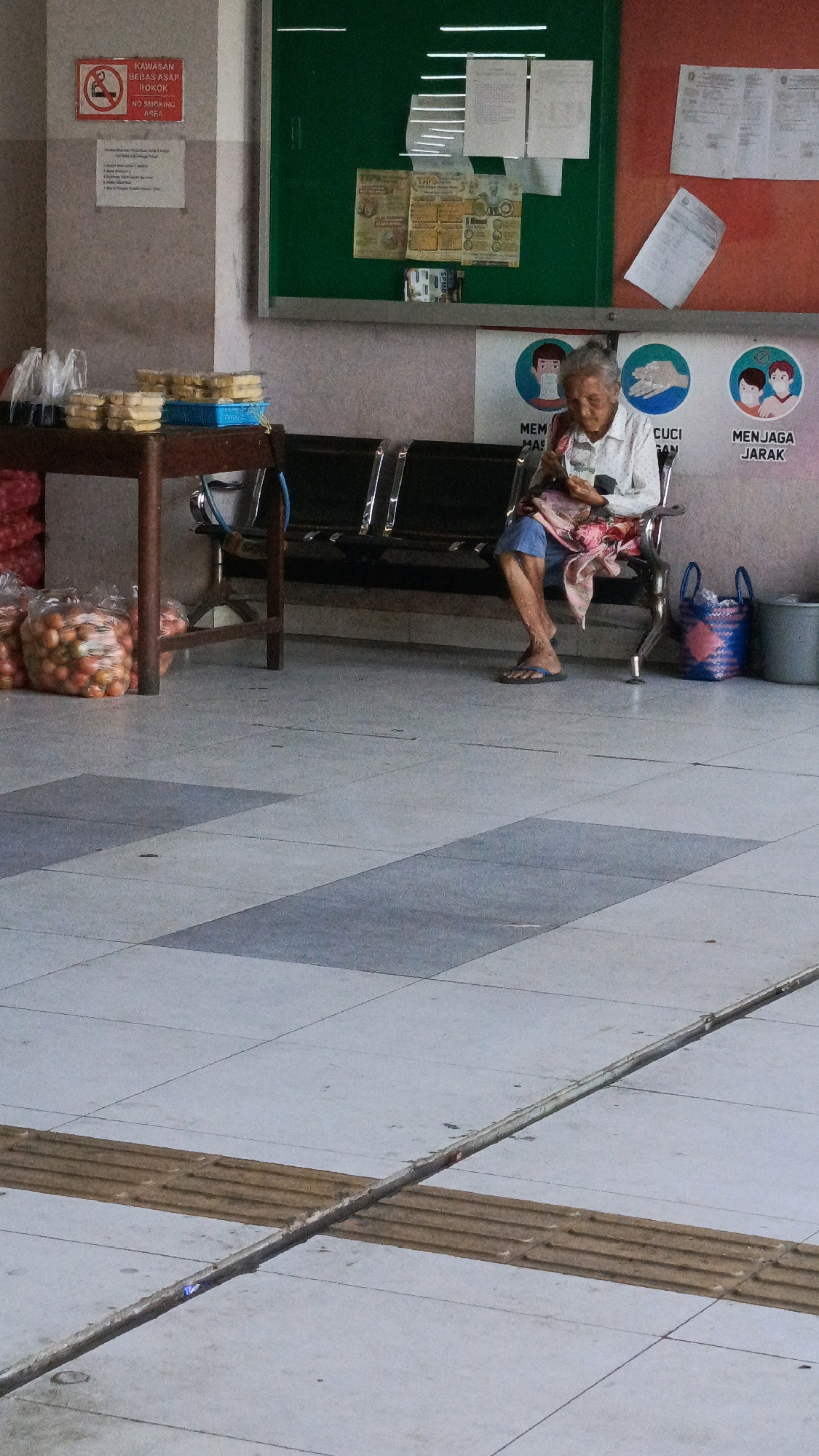An elderly woman is seated on a metal bench in what appears to be a waiting area, possibly at a public facility or market, in Ponorogo, Indonesia. She is wearing a light-colored patterned shirt and blue shorts, with a pink cloth draped over her lap. She is looking down at something in her hands. In front of the bench, there are several bags of produce, likely tomatoes, and a table with packaged food items. The floor is tiled with a gray and blue pattern. To the right of the woman, a blue and pink woven basket and a gray trash can are visible. Behind the bench, there are posters with health advisories, including messages about wearing masks, washing hands, and social distancing, indicated by icons of people wearing masks and washing hands. The overall environment suggests a public space with ongoing health awareness campaigns.