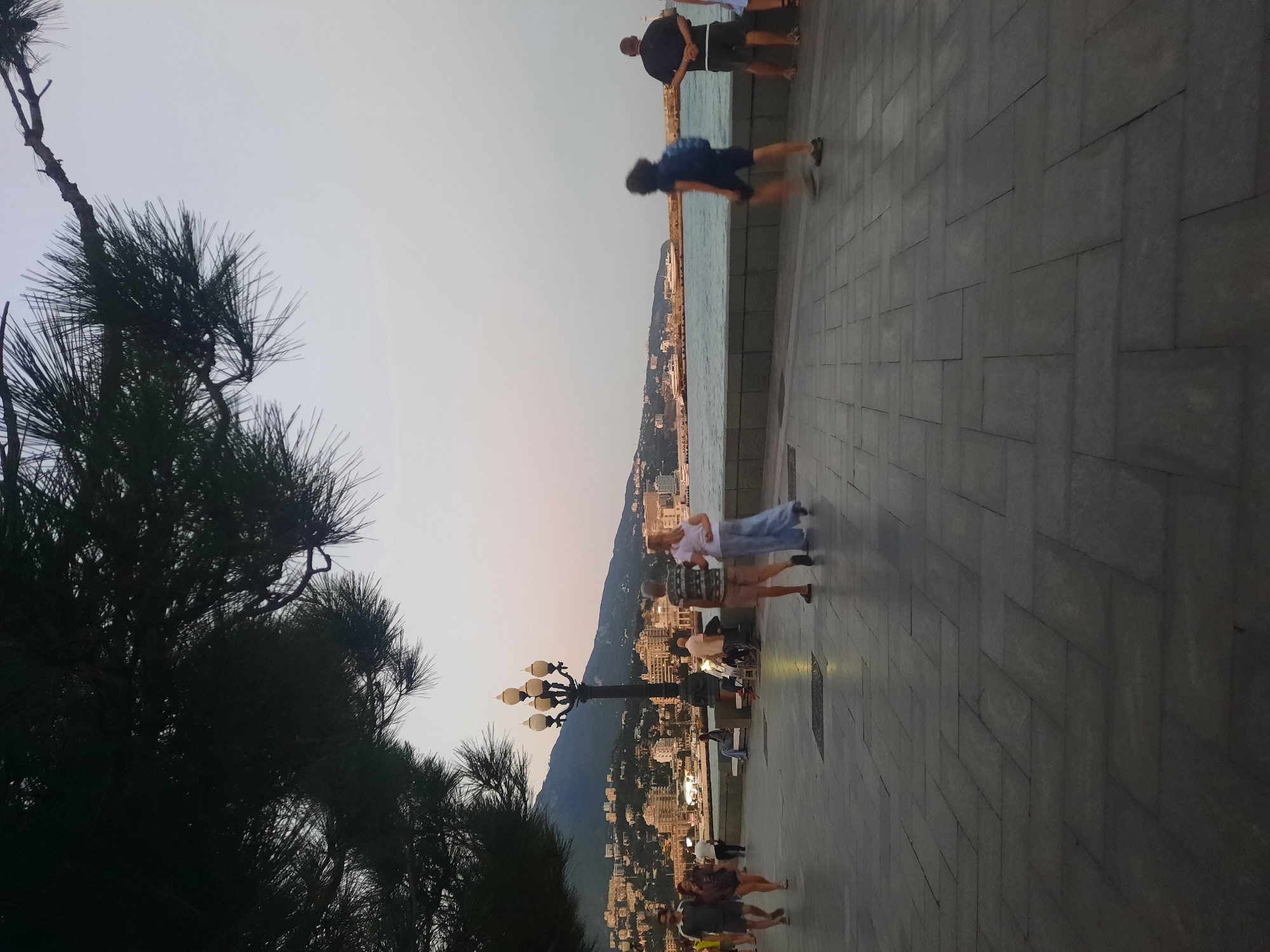 The image depicts a seaside promenade in Yalta, Ukraine, during the late afternoon or early evening. A large pine tree with dark green needles partially obscures the top left of the frame. In the middle ground, a multi-headed lamppost stands on the paved walkway. Several people are visible strolling along the promenade. To the left, a couple walks away from the camera, with the man in a grey shirt and dark shorts, and the woman in a dark top and shorts. Further along, another couple walks towards the camera; the man wears a patterned t-shirt and beige shorts, and the woman wears a white t-shirt and light blue trousers. In the distance, a blurry figure with a blue backpack walks towards the right of the frame, and to their right, a man in a black t-shirt and shorts stands with his hands behind his back. The background features the sea, with several ships visible on the water. Beyond the sea, a hillside city with numerous buildings and lights stretches along the coast, leading up to a dark mountain range under a pale, hazy sky. The promenade itself is paved with large, rectangular grey stones laid in a herringbone pattern.