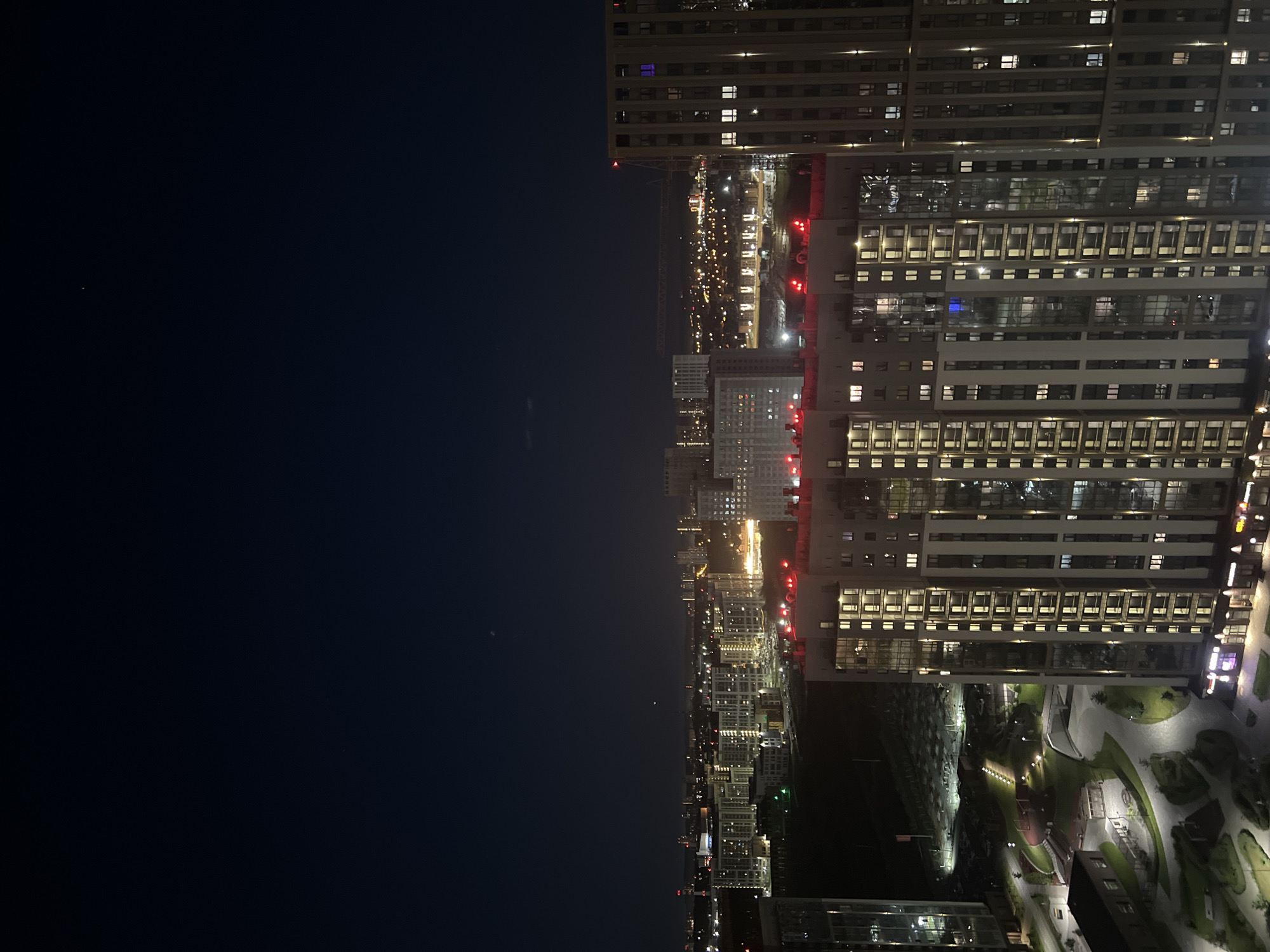 The image captures a nighttime aerial view of a modern urban landscape in Yekaterinburg, Russia. The foreground is dominated by two tall residential buildings, characterized by their illuminated windows that create a grid pattern against the dark facade. The building on the right is taller and wider, with visible vertical accent lighting. The building on the left, partially visible, appears to be a slender high-rise.
Below the foreground buildings, a structured outdoor area is visible, featuring pathways, green spaces, and playground equipment, all illuminated by low-level lighting. Further in the distance, a sprawling cityscape unfolds, with numerous buildings of varying heights illuminated by a multitude of lights. Some buildings have red lights on their rooftops, possibly indicating helipads or safety markers. A faint glow from distant lights suggests a broader urban expanse. The sky above is a deep, dark blue, with a few faint celestial bodies visible. The overall impression is one of a well-lit, modern residential district at night.
