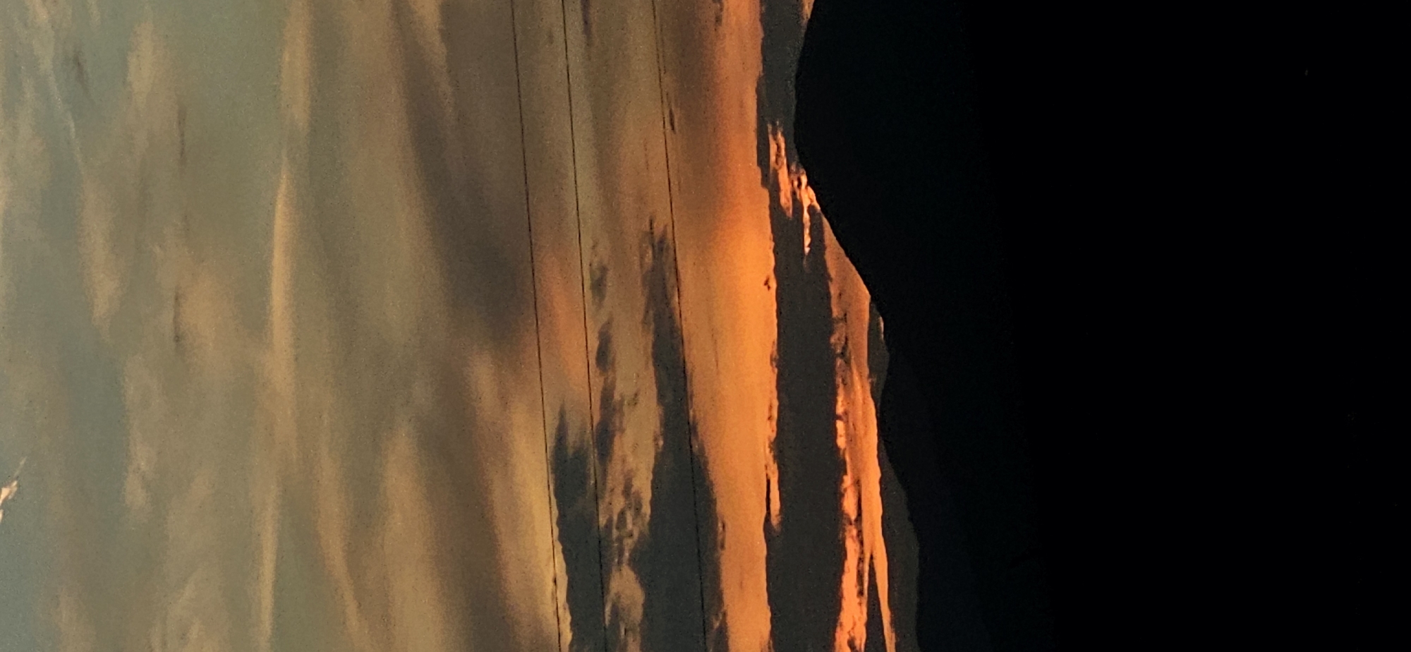A nighttime photograph captures a silhouette of mountains against a vibrant sunset sky. The sky is a gradient of oranges and blues, with streaks of clouds running horizontally across the frame. Power lines are visible in the upper portion of the image, spanning the sky. The foreground is dominated by the dark, shadowy shapes of rolling hills or mountains, with the crest of one larger peak visible on the right side. The scene appears to be an outdoor landscape, likely in a rural or mountainous area. This location is identified as being in Momin Prohod, Bulgaria.