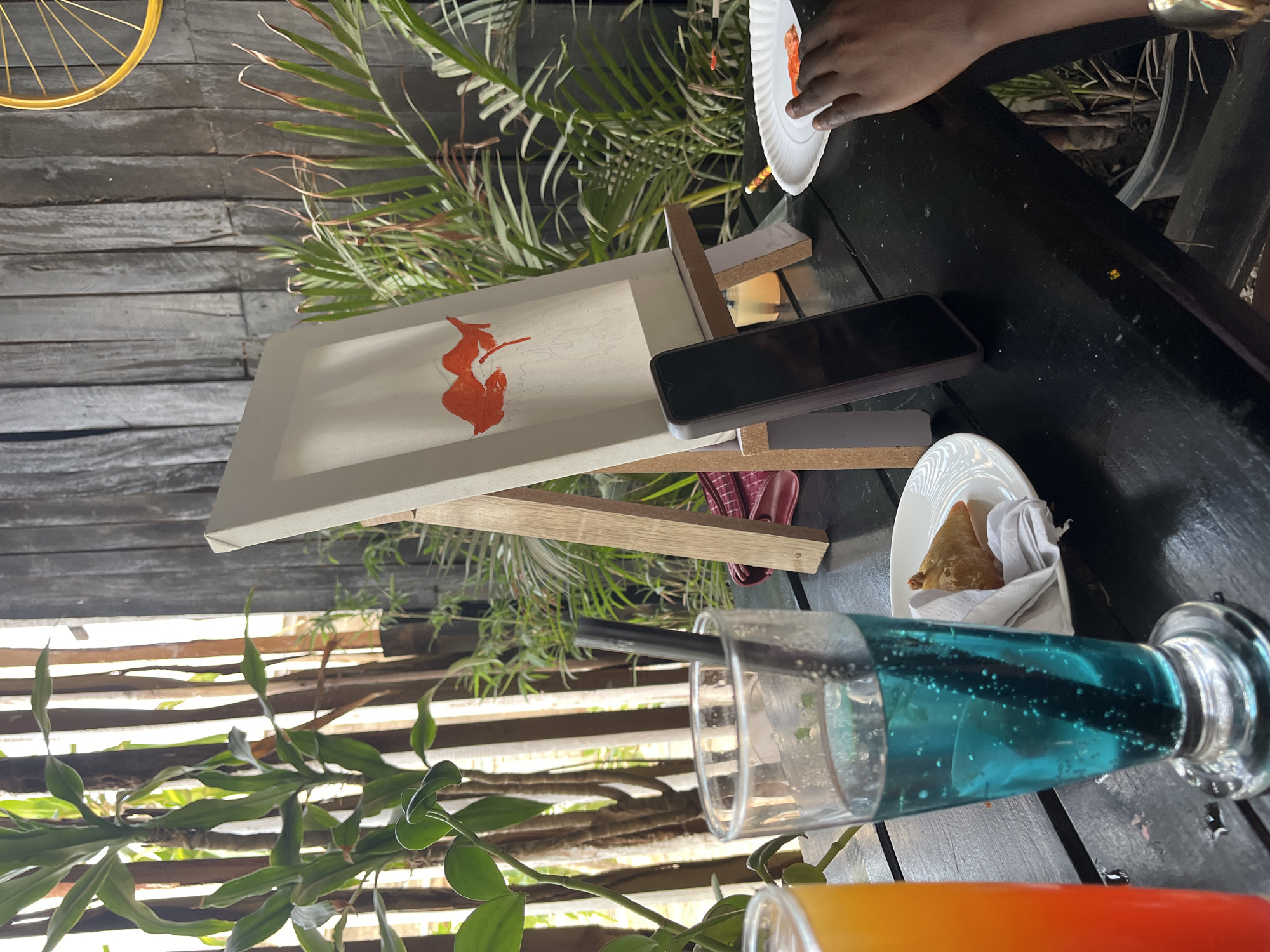 On a dark, seemingly wooden table, possibly outdoors, various objects are arranged. A painting on an easel with an orange abstract design takes center stage, a black smartphone rests on the easel's base. A plate holding a triangular pastry and some white napkins sits beside the painting. Two glasses are visible: one filled with a blue liquid and the other with an orange liquid, with a black straw in the blue drink. A hand reaches for food on a white plate. The background features greenery and what appears to be a grey slatted structure. Given the specific request, it's impossible to verify definitively the location in Jos, Nigeria, without further information.