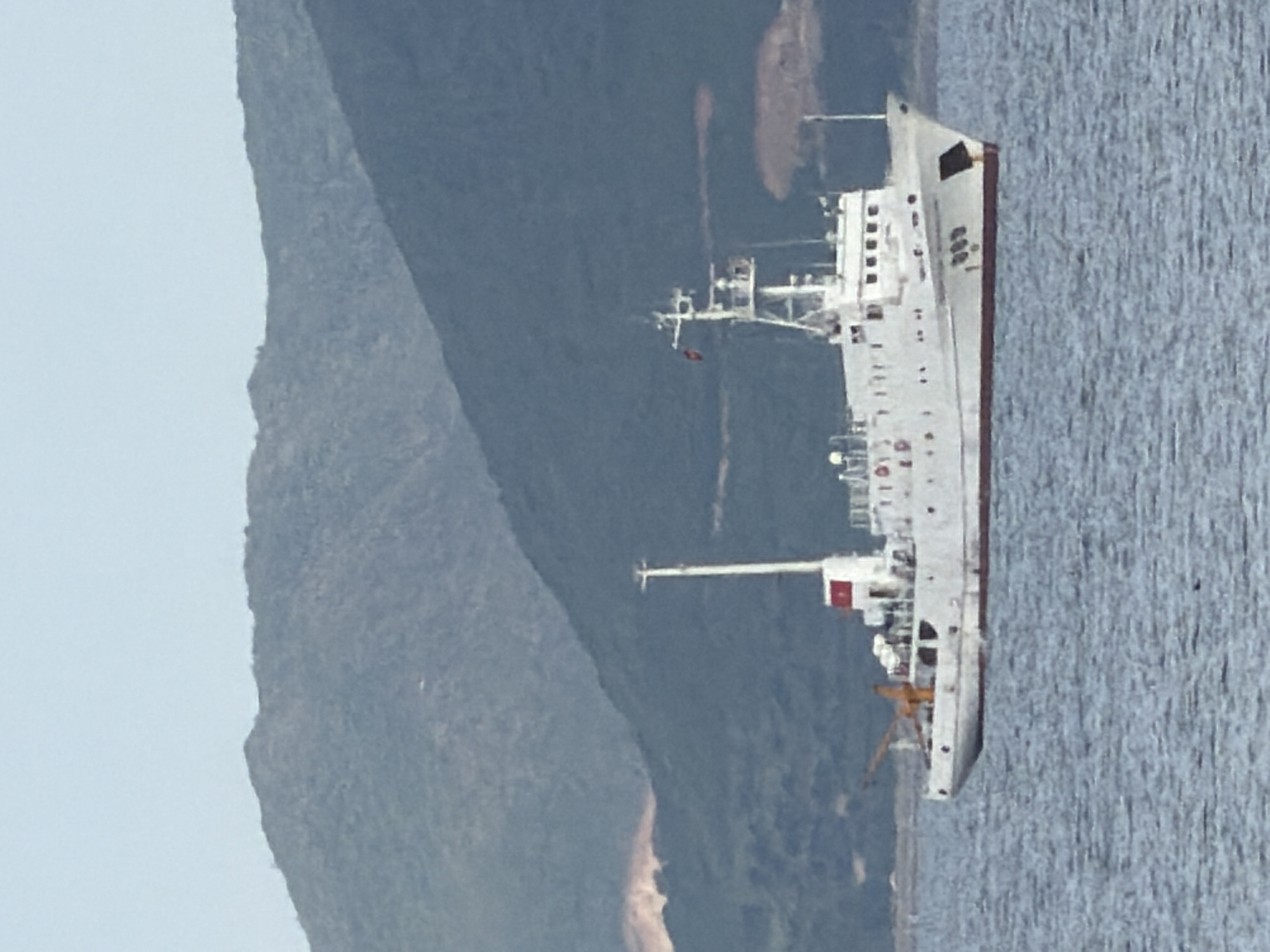 A white ship with a red waterline is sailing on the water. The ship has a tall mast with a flag and several windows. Behind the ship, there are mountains and a blue sky. The water is rippled.