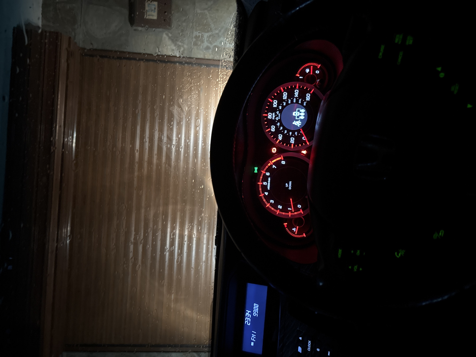 The image depicts the interior of a car, specifically the dashboard and instrument panel, viewed through the windshield on a rainy night. The speedometer reads approximately 48 mph, and the radio is tuned to FM 99.00 at 23:34. Outside, the view through the windshield shows a closed brown roll-up door. The location is unknown. There are no people visible.