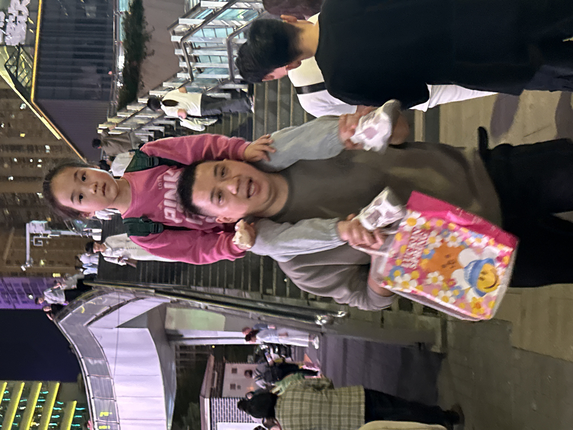 This image, likely captured in Yuzhong District, China, depicts a father carrying a young girl on his shoulders, facing the camera. The man, with short dark hair, wears a grey or olive-colored long-sleeved top and dark pants, and has an expressive, open-mouthed look, possibly smiling or talking. The girl, positioned on his shoulders, is dressed in a pink long-sleeved shirt with the word "PINK" visibly printed on it. She has dark hair and a neutral to slightly curious expression as she looks towards the viewer.
The man is holding a clear plastic bag in his left hand, which features a cheerful design of white and yellow flowers and a cartoon character resembling a bear in a blue hat, along with some illegible text.
The scene is set outdoors in an urban environment during what appears to be the evening or night, indicated by the dark sky and reliance on artificial lighting. In the background, numerous people are visible on a multi-level pedestrian area, consisting of stairs, escalators, and walkways with modern metal railings. The architecture suggests a contemporary public space or shopping district. Some faint yellow and green lights from distant buildings or street lamps are visible in the far background. The overall atmosphere appears lively and bustling.