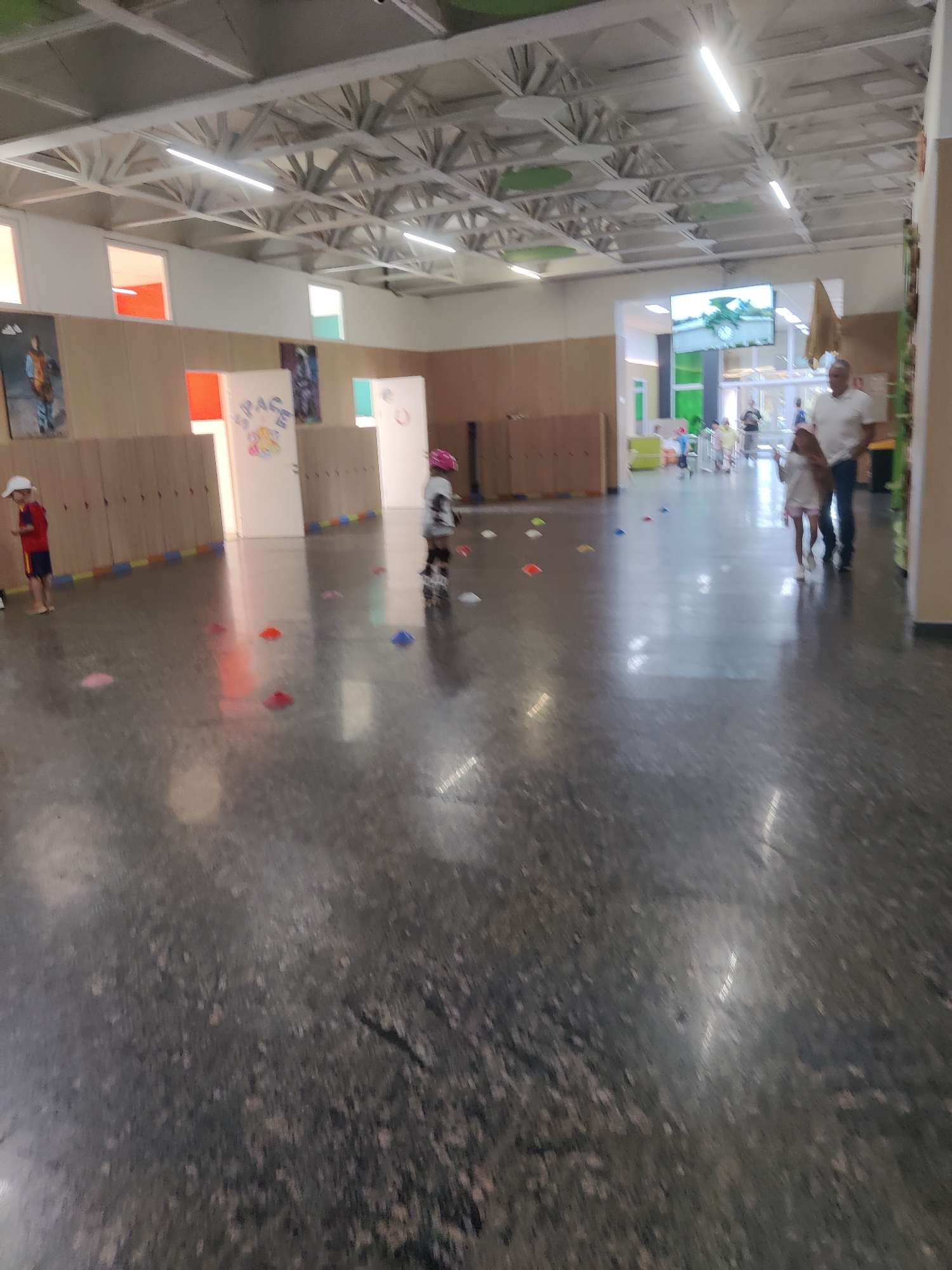 The image depicts an indoor space, possibly a gymnasium or multi-purpose room, with a high ceiling featuring exposed beams and fluorescent lighting. The floor is a dark, speckled material, and several colorful cones are scattered across it, suggesting an activity or obstacle course.
On the left side of the image, a young child wearing a red shirt and blue shorts stands with a white hat, facing away from the camera. Further into the room, another child on roller skates, wearing a helmet and protective gear, is also visible.
In the background, an adult man in a white polo shirt and dark pants is walking with a young girl. Other children and adults are present in the distance, moving through the space. The walls are lined with light-colored wooden cabinets, and there are several doors and windows. Artwork or posters are visible on some of the doors. The overall atmosphere suggests a children's activity or event.
The location is identified as being in the city of Kyiv, Ukraine.
