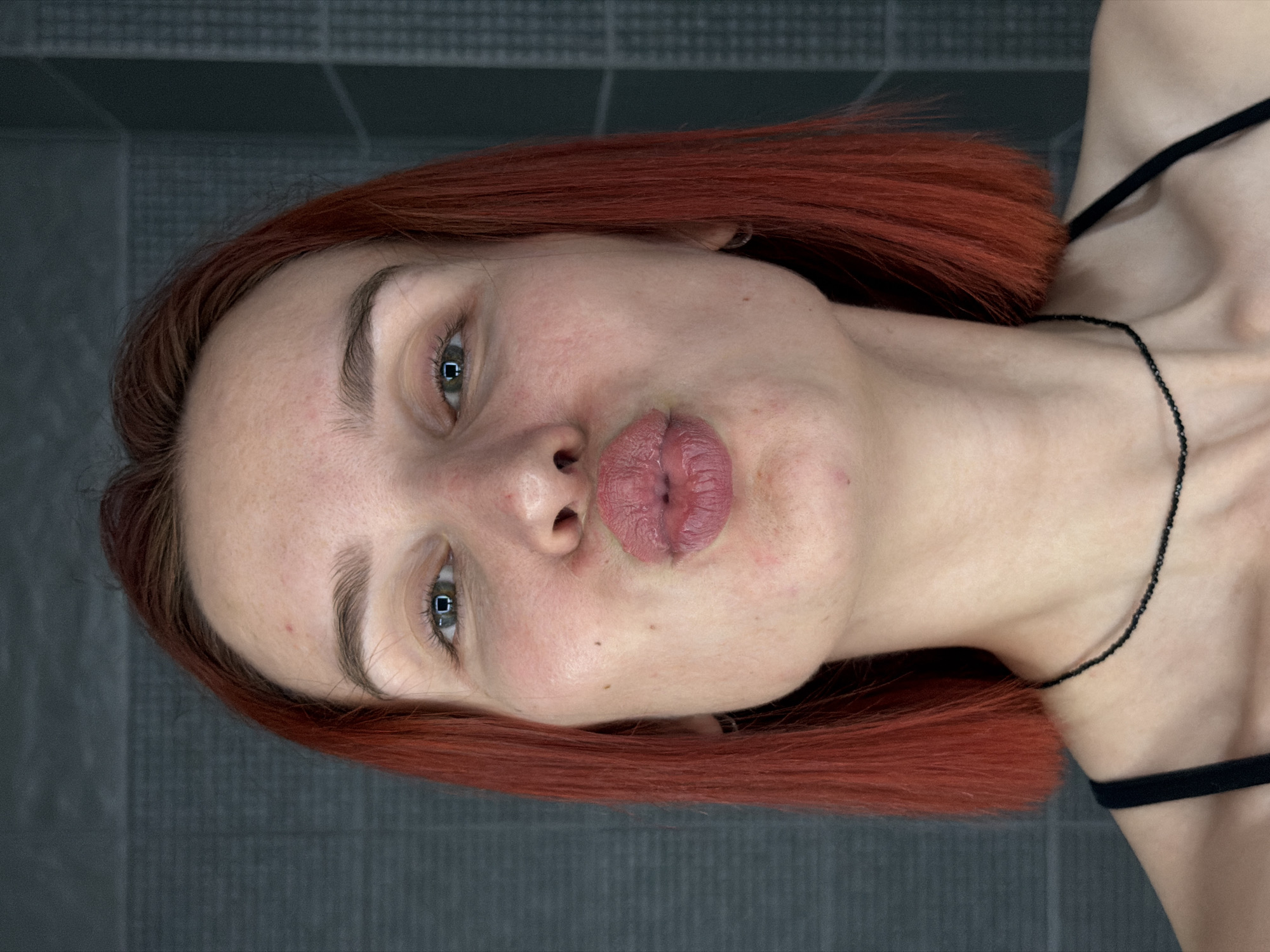 A close-up, tilted portrait shows a fair-skinned woman with green eyes and pursed lips, possibly in a kissing gesture. She has short, reddish-brown hair and a dark necklace. A black tank top strap is visible on her right shoulder. The background is a gray tiled surface. The location is not discernible from this image and cannot be confirmed as Chaikovsky, Russia.