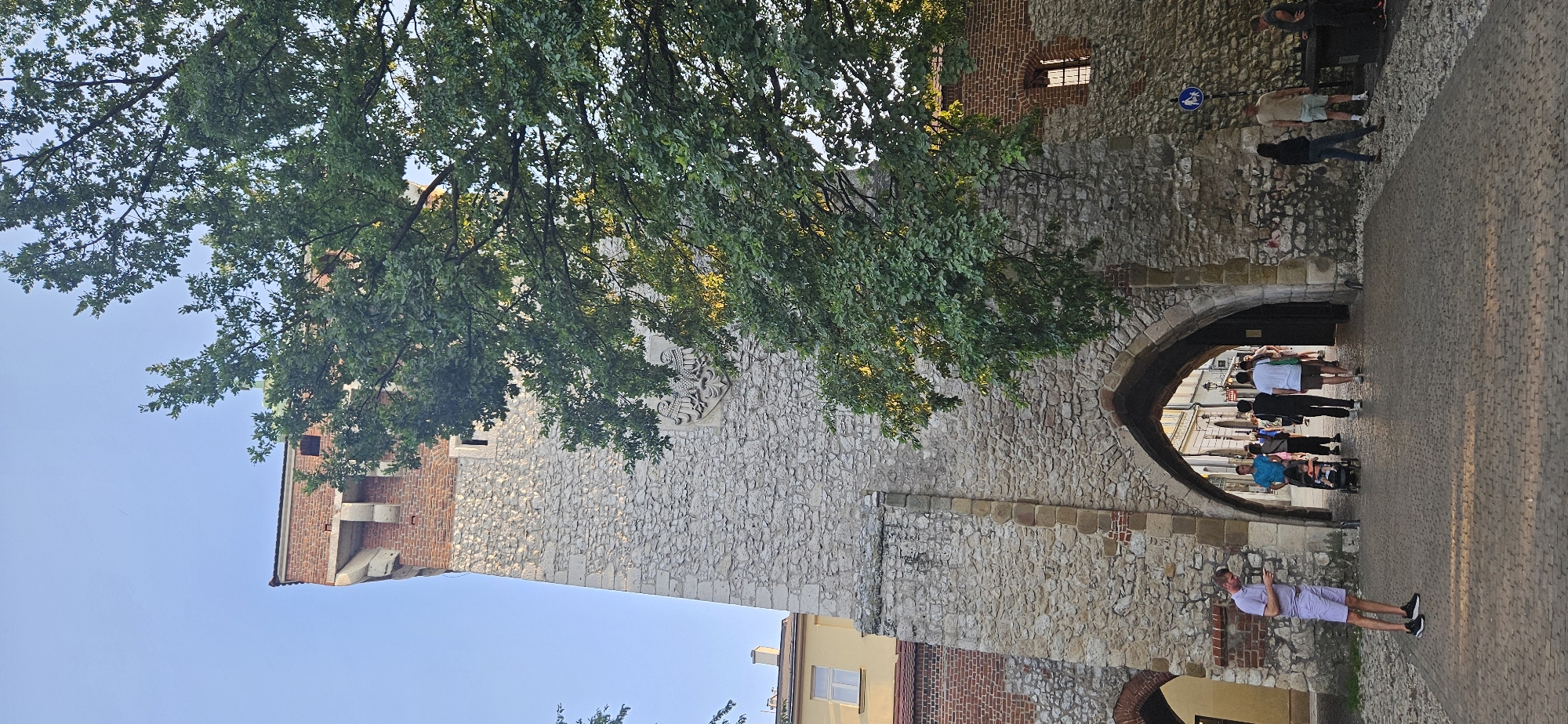 In Krakow, Poland, a stone tower with arched gateways stands tall, partially obscured by a large tree with green leaves. The tower's lower section is constructed from rough-hewn stones, while the upper portion is made of red brick. An ornate stone crest is visible on the tower's face. Several people are walking through the archways, some with shopping bags and one pushing a stroller. The ground is paved with cobblestones, and a blue sign with white figures indicating pedestrian crossing is posted on the wall to the right of the archway. The sky is clear and blue.