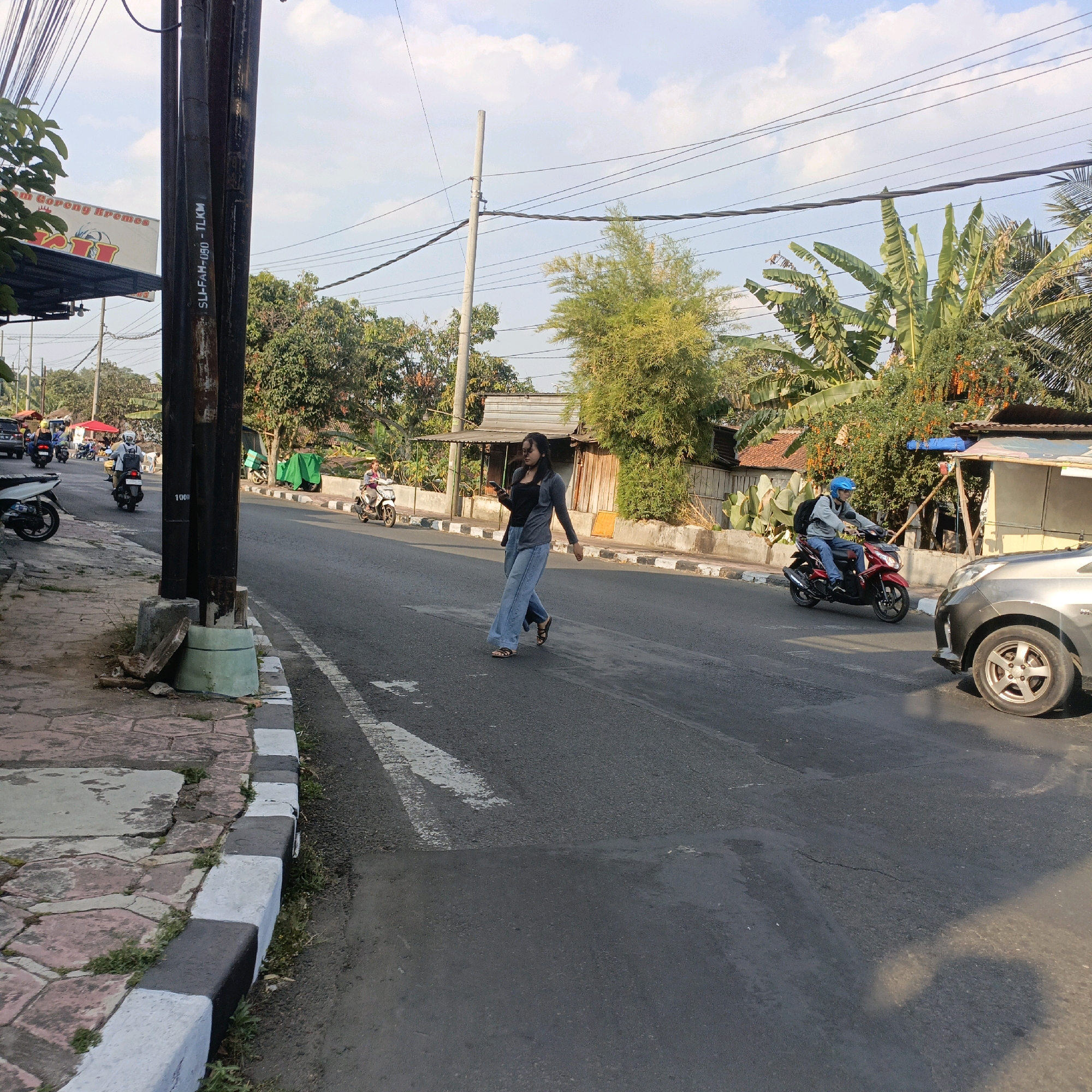 A young woman is walking across a street in Salatiga, Indonesia, looking at her phone. She is wearing a grey cardigan over a black top and wide-leg blue jeans. She is wearing dark sandals and has dark hair. The street is paved and has a crosswalk. There are several motorcycles and cars on the street, and a few pedestrians on the sidewalk. The background shows a line of trees and buildings, with power lines overhead. The sky is blue with some clouds. The scene appears to be a typical street scene in a city in Indonesia.