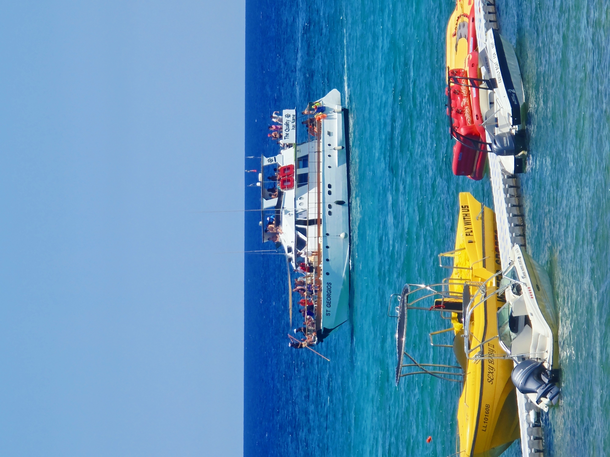 The image depicts a sunny day at sea, likely in the city of Protaras, Cyprus, given the boats and clear blue waters. In the background, a white passenger boat named "ST GEORGIOS" is visible, carrying a group of people on its decks. The boat is navigating through turquoise waters, with more people on the upper deck enjoying the ride. Text on the boat reads "The Quality has Name."
In the foreground, several smaller boats are docked at a floating pier. These include a yellow inflatable boat with the text "SEXY BABY II" and registration "LL10160B" on its side, and another yellow inflatable boat with text "FLY WITH US". Beside these, a white motorboat with a blue stripe is docked, featuring the text "FAMILY BOAT FOR RENT" and a Yamaha engine. A red inflatable watercraft is also visible behind the yellow boats. The scene is characterized by bright sunlight, clear blue skies, and calm, vibrant blue ocean waters, suggesting a typical recreational day at a coastal resort.