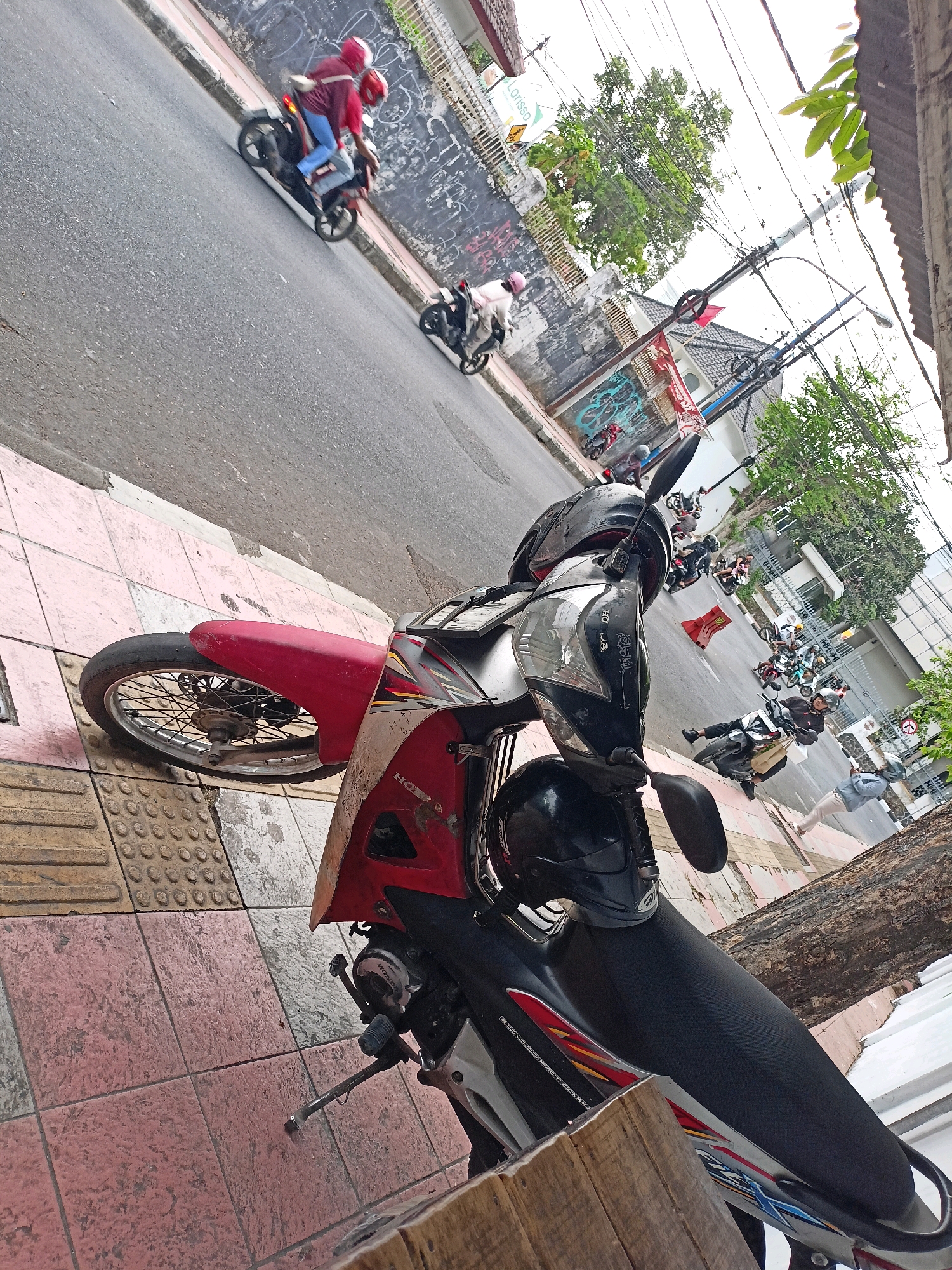 The image captures a street scene in Magelang, Indonesia, from a tilted perspective. In the foreground, a red and black motorcycle is parked on a tiled sidewalk. The motorcycle has a black helmet resting on its front. Beyond the sidewalk, the street is visible with several other motorcycles, some carrying passengers, in motion. A few individuals are also seen walking or standing on the sidewalk. The background shows buildings, trees, utility poles with numerous wires, and a concrete wall with graffiti. The overall atmosphere suggests a typical day in a busy urban environment.