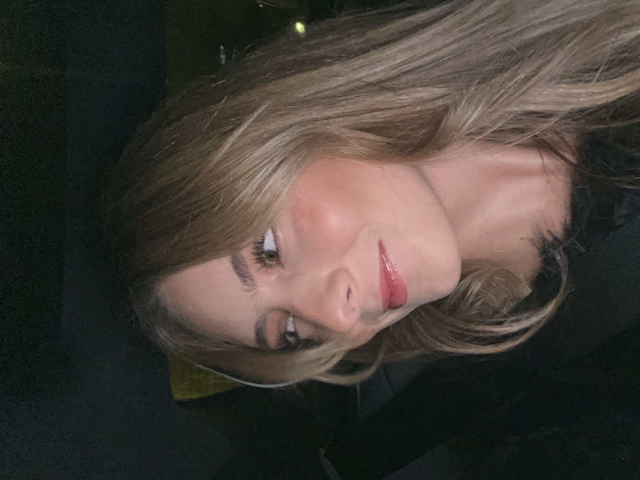 A young woman with blonde hair looks toward the camera with a slight smile. She has full lips with a glossy pink lipstick, prominent cheekbones, and long eyelashes. She appears to be in a car, as evidenced by the dark interior and seatbelt visible in the background. Her hair is styled with layers, and her complexion is fair. She is wearing a dark top with some kind of soft texture visible around the neckline.