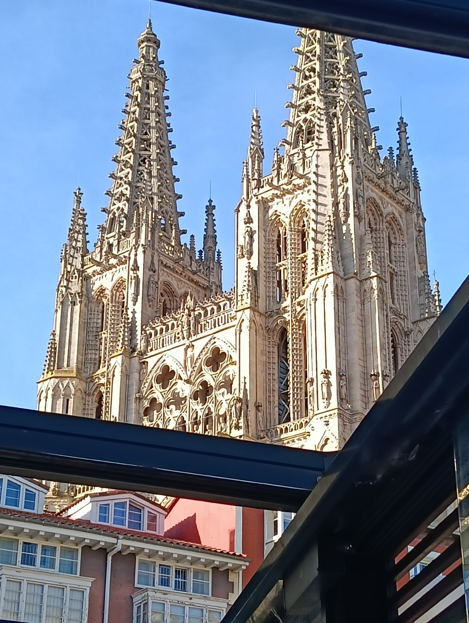 The image shows the Burgos Cathedral, a prominent example of Gothic architecture in Burgos, Spain. The cathedral's two spired towers are prominently featured, showcasing intricate stonework and detailing against a clear blue sky. The photo is taken from a slightly lower angle, with a modern metal structure visible in the foreground, partially framing the view of the cathedral and nearby buildings. Lower buildings with red roofs and white window frames can be seen below the cathedral. No people are visible in the scene.