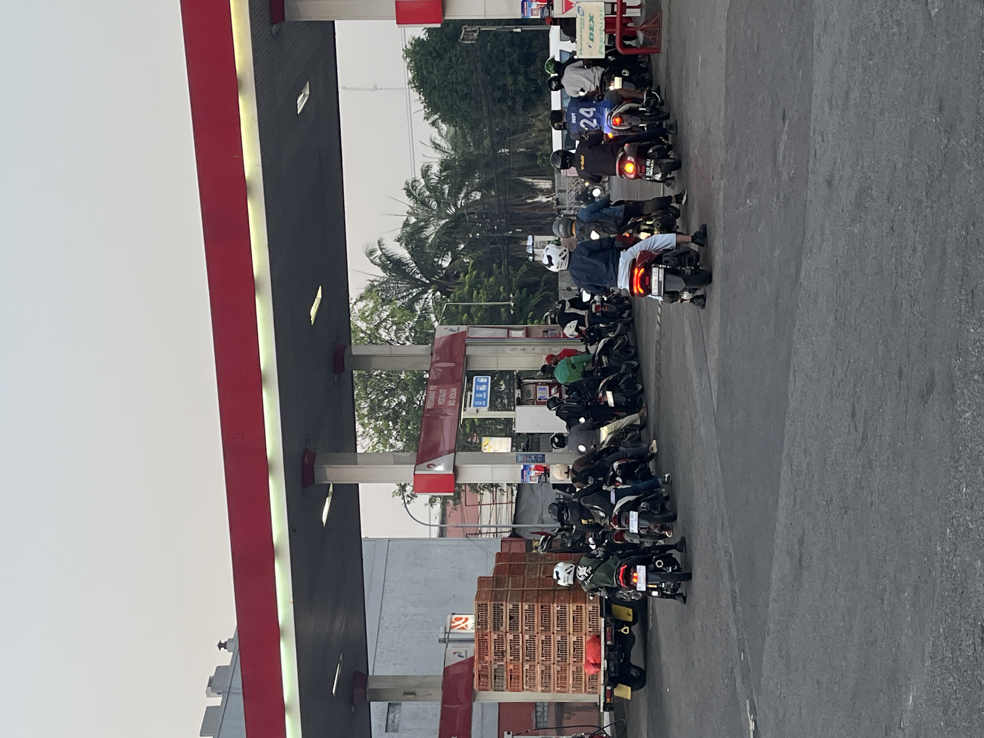 This is a medium shot, eye-level, vertical photograph of a busy gas station in Tambun, Indonesia. The scene depicts a long line of motorcycles and a truck waiting to refuel. The gas station canopy, painted in red and dark gray, dominates the upper portion of the image, with bright fluorescent lights illuminating the area. Several fuel pumps are visible, with signage indicating different fuel types.

In the foreground, a flatbed truck is parked, loaded with stacks of wooden crates. A queue of motorcycles, ridden by individuals wearing helmets, stretches from the truck towards the fuel pumps. The riders are mostly facing away from the camera, indicating they are waiting their turn. The road surface is asphalt.

In the mid-ground, more motorcycles and their riders are lined up. Some individuals are standing next to their bikes. The background features a building with a gray facade, palm trees, and other greenery, suggesting a tropical or subtropical environment. The sky is overcast and pale, typical of an early morning or late afternoon. The overall atmosphere is one of routine activity at a commercial establishment.