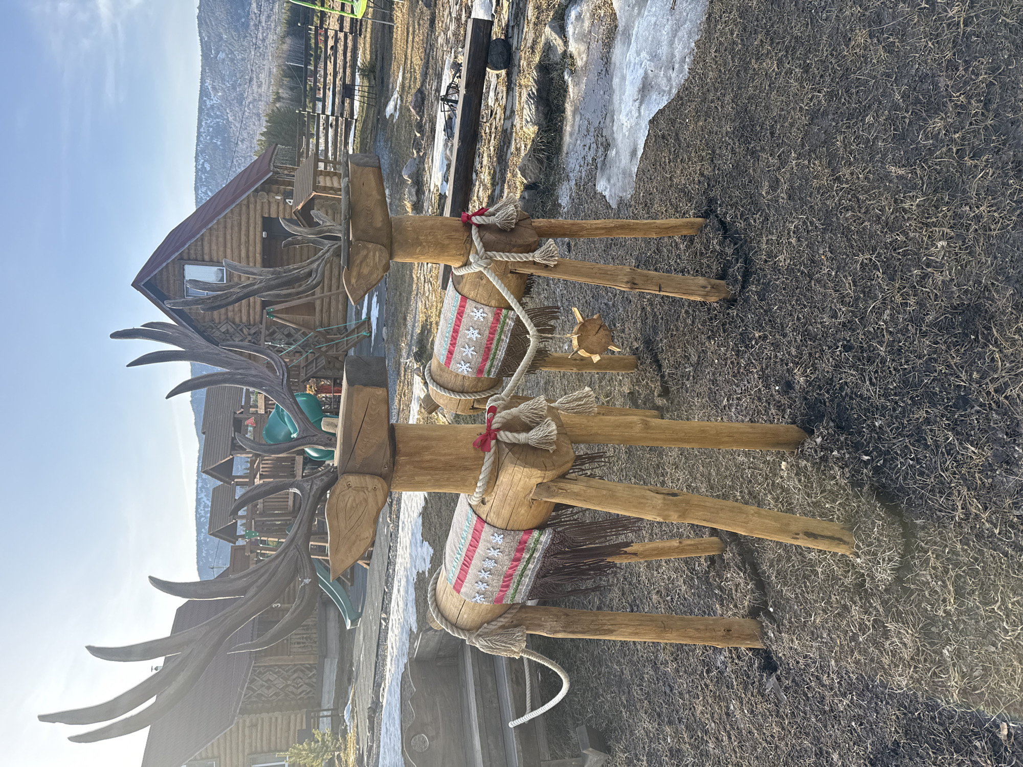 Two life-sized wooden reindeer sculptures stand in the foreground of an outdoor scene in Tyulyuk, Russia. Each reindeer is constructed from logs and branches, featuring carved heads with multi-pronged antlers. Their cylindrical bodies are adorned with woven blankets displaying geometric patterns in red, green, white, and gray, accented with white snowflake motifs. Ropes with braided tassels are secured around their midsections, simulating harnesses. The leading reindeer has a small, star-shaped wooden charm suspended from its rope. The trailing reindeer is positioned to the left and slightly behind, its rope extending towards a rustic wooden sleigh or bench.
The ground is dark, damp earth covered with sparse, dry grass. Patches of melting snow and ice are visible, particularly towards the right side and in the middle distance. In the background, several log cabins with red gable roofs are visible, along with a multi-level wooden playground featuring green slides. A wooden fence delineates a portion of the property. Beyond these structures, a natural landscape unfolds, consisting of evergreen trees and snow-capped mountains under a clear blue sky. Sunlight from the upper left casts distinct shadows forward and to the right of the reindeer sculptures.