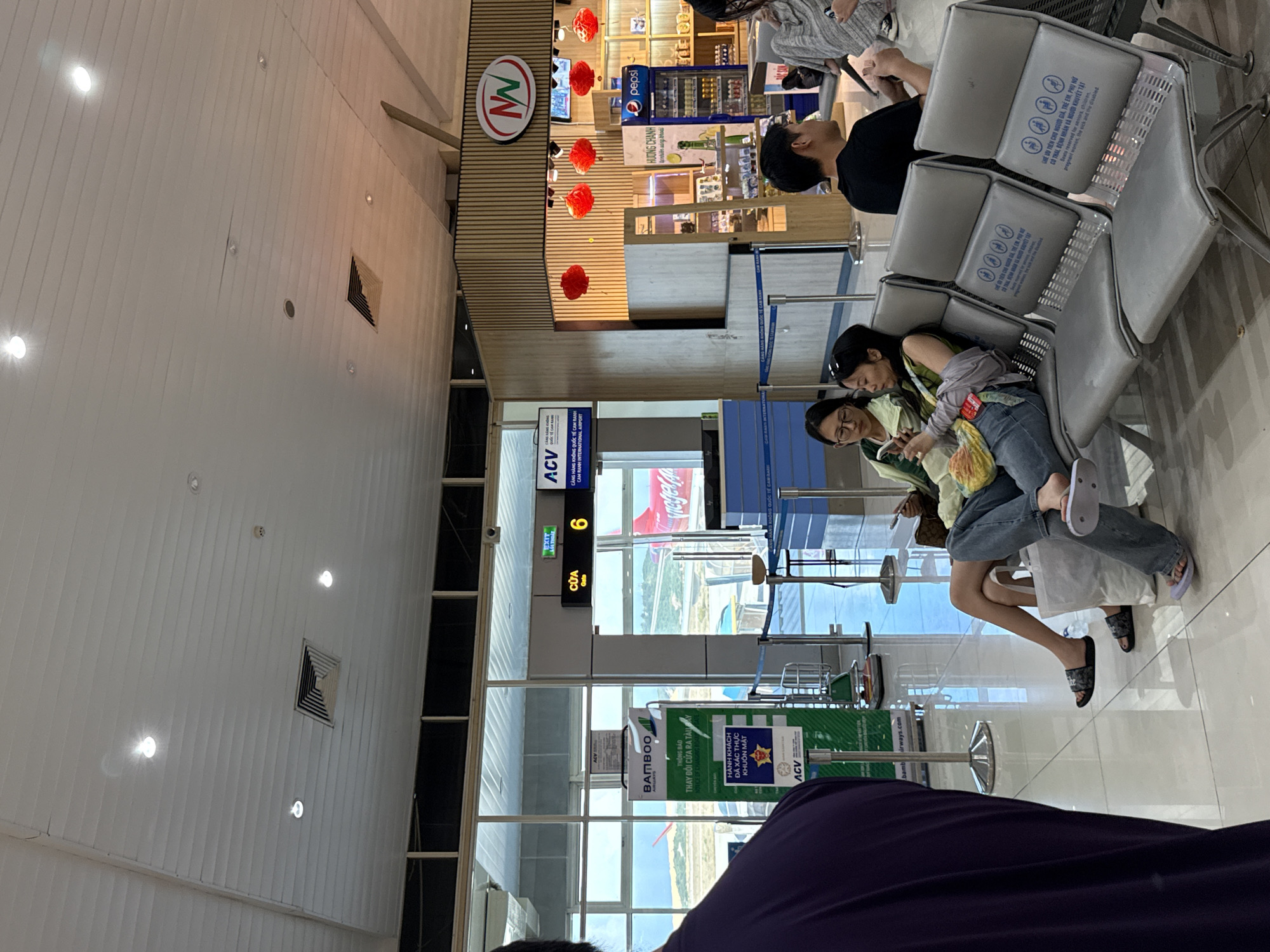 The image shows the interior of Cam Ranh International Airport in Bac Cam Ranh Ward, Vietnam. Two women are seated in metal chairs, looking at their phones. Another man is standing by the seats, also looking at a phone. Behind the women and man are an airport gate, with signage indicating Bamboo Airways, gate number 6, and ACV (Airports Corporation of Vietnam). There's also a snack shop in the background selling beverages such as Pepsi and "Huong Chanh".